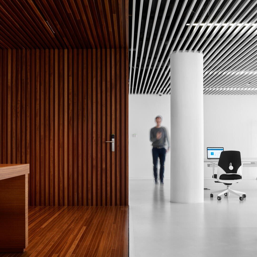 Ein moderner Büroraum, geteilt durch eine Holzwand auf der linken Seite und eine weiße Säule auf der rechten Seite, mit einer unscharfen Person beim Gehen, einem weißen Schreibtisch und einem Computer in einer hellen, minimalistischen Umgebung.