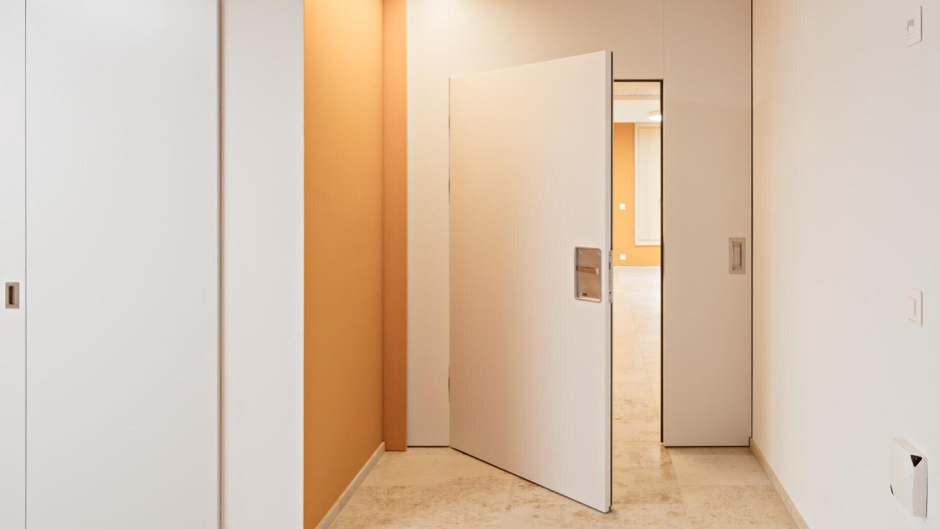 Un couloir intérieur moderne avec des sols beiges, des murs blancs, une porte coulissante sur la gauche et une porte battante blanche qui s'ouvre partiellement pour laisser apparaître une pièce aux murs jaune-orange chaleureux.