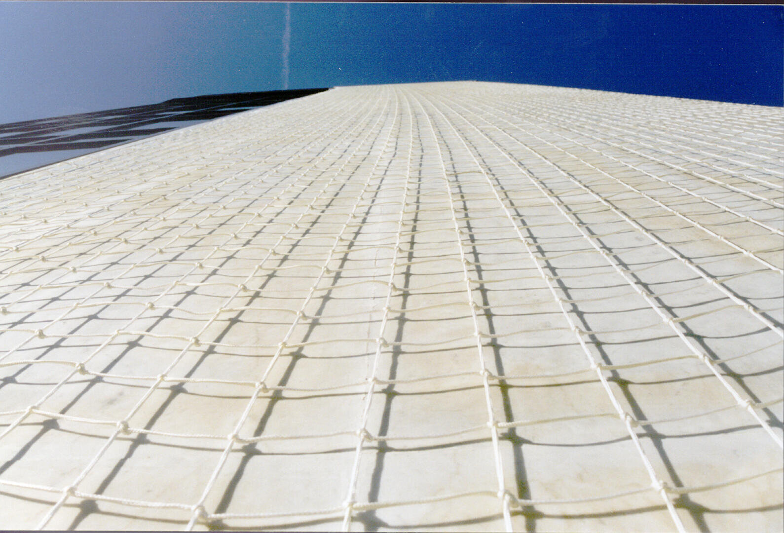 Vue à basse altitude d'une haute structure recouverte d'un filet blanc à motif de grille, sur fond de ciel bleu clair. La perspective souligne la hauteur et les lignes verticales de la structure.