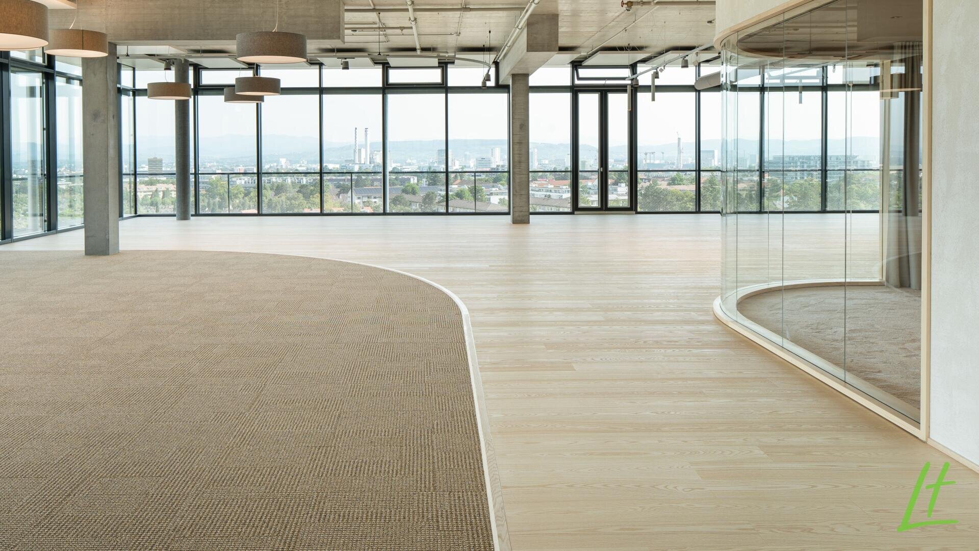 Espace de bureau moderne et vide avec de grandes fenêtres, une vue sur la ville, des sols en bois et en moquette, des plafonniers et une paroi vitrée incurvée sur le côté droit. La lumière naturelle inonde l'espace ouvert et spacieux.