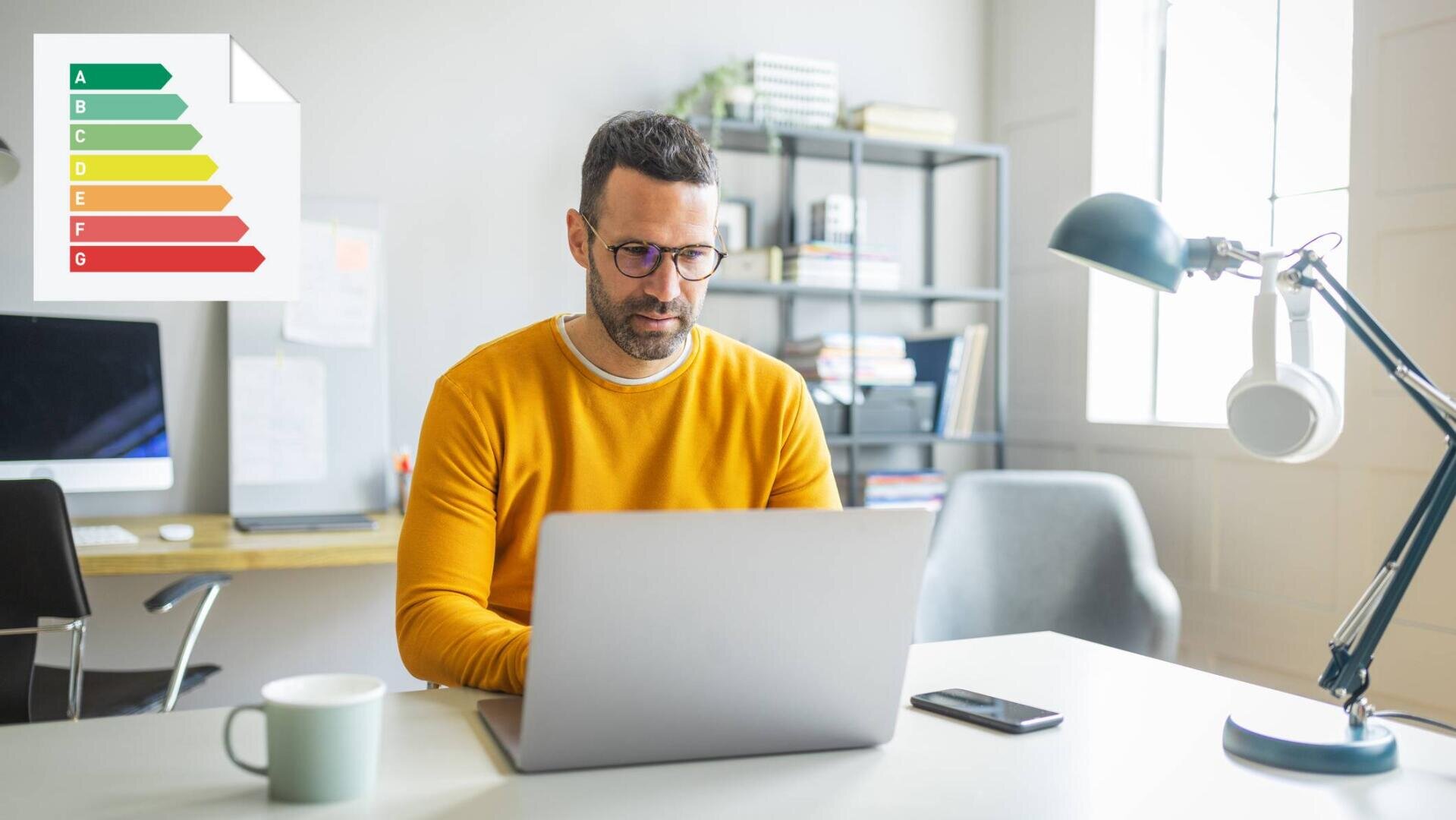 Ein Mann mit einer Brille und einem gelben Pullover arbeitet an einem Laptop an einem Schreibtisch. Auf dem Schreibtisch stehen ein Kaffeebecher, ein Smartphone und eine Lampe. An der Wand hinter ihm ist eine Tabelle mit Energieeffizienzwerten zu sehen.