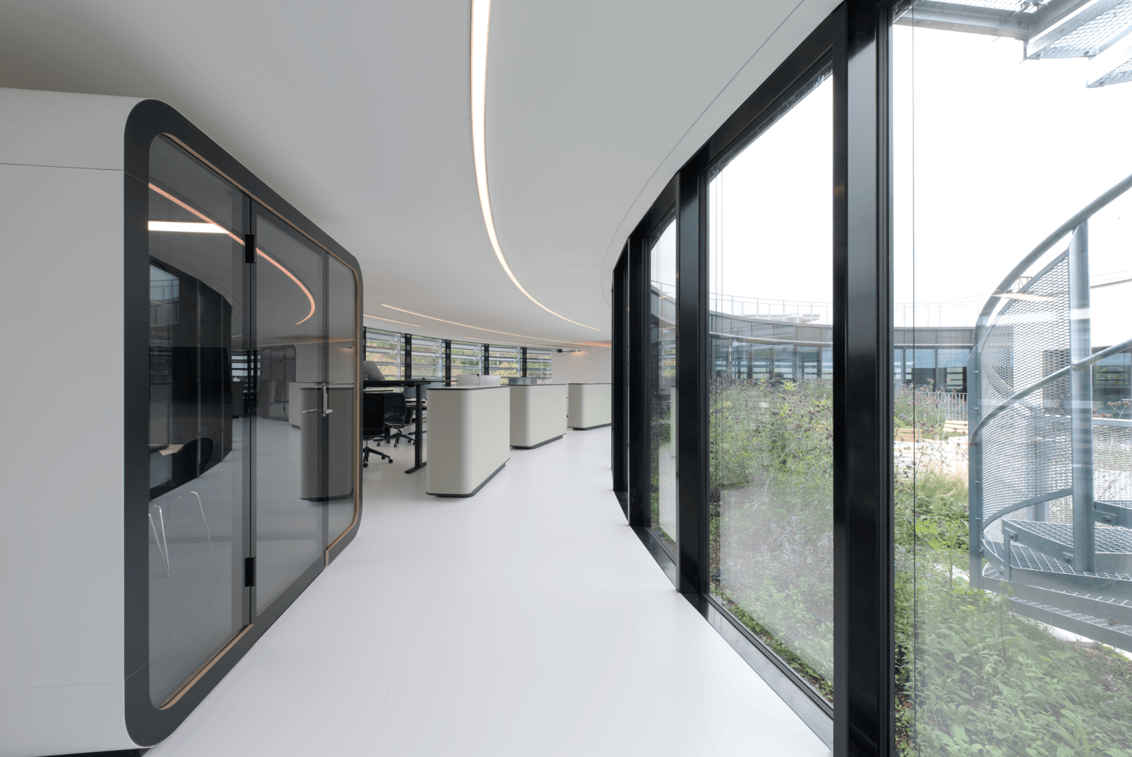 Modernes Bürointerieur mit geschwungenen Glaswänden, großen Fenstern mit Blick ins Grüne, minimalistischen weißen Böden und linksseitig geschlossenen Besprechungsräumen. Natürliches Licht füllt den offenen und geräumigen Arbeitsbereich.
