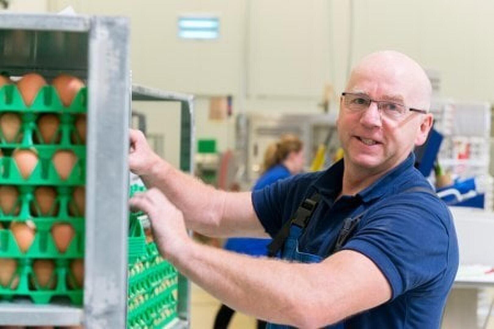 Ein Mann in blauem Hemd und Schürze steht in einer Fabrik oder einem Lagerhaus neben den Regalen mit Eiern und lächelt in die Kamera, während er mit den Schalen arbeitet.