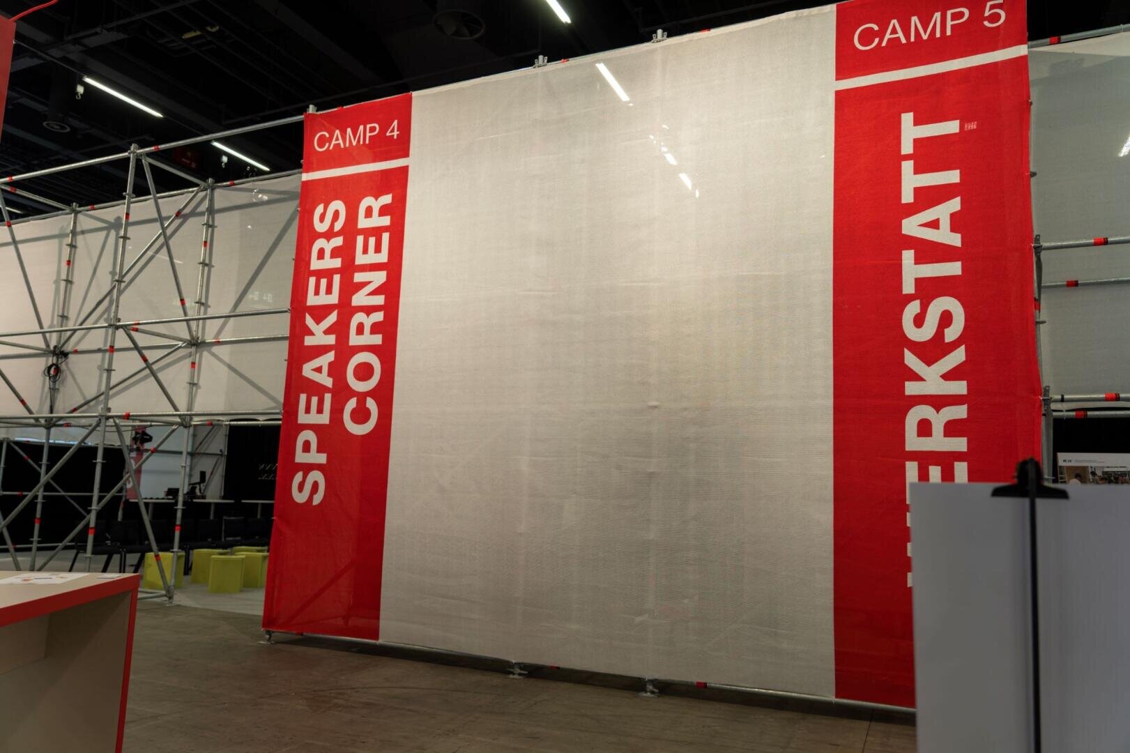 Une grande cloison rouge et blanche sur un lieu d'événement affiche les mots SPEAKERS CORNER et WERKSTATT avec l'inscription CAMP 4 et CAMP 5 sur le bord supérieur, à côté d'une structure métallique.