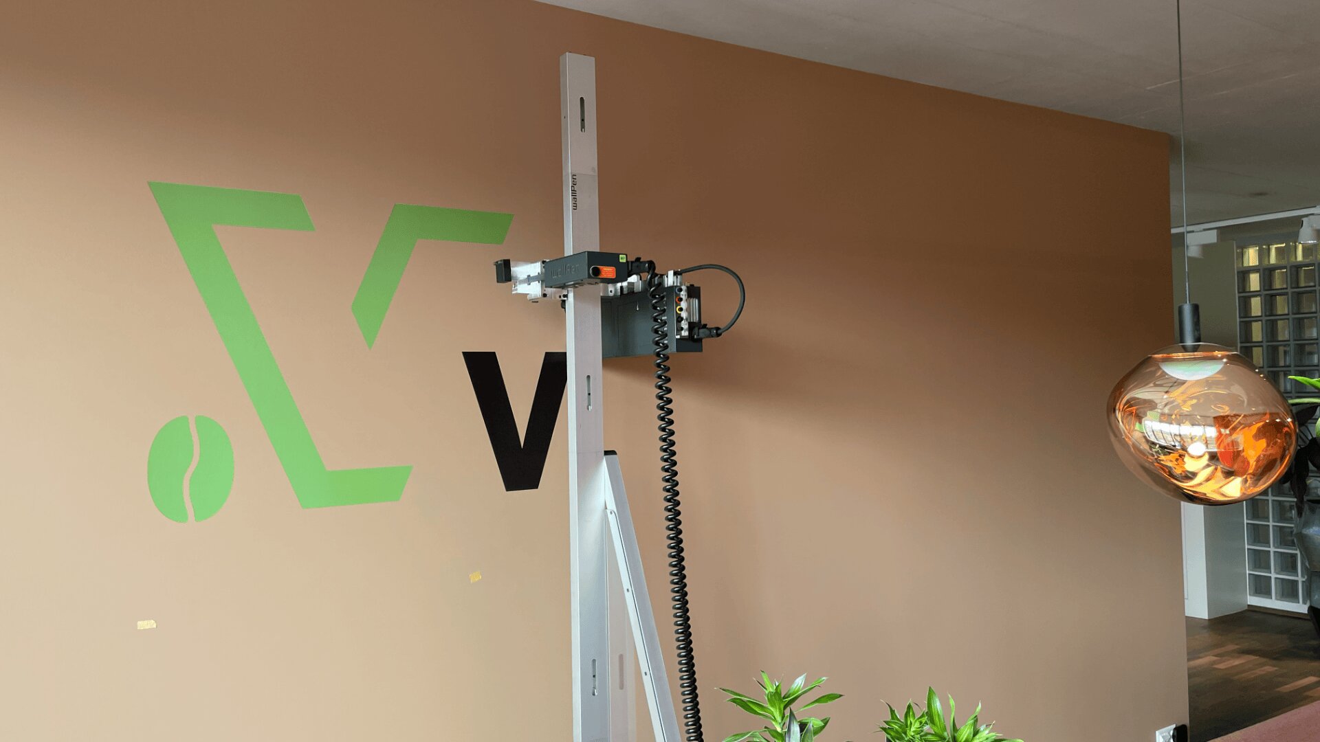 Une imprimante murale imprime un logo vert et noir sur un mur brun dans une pièce moderne avec une suspension ronde et des plantes en pot à proximité.