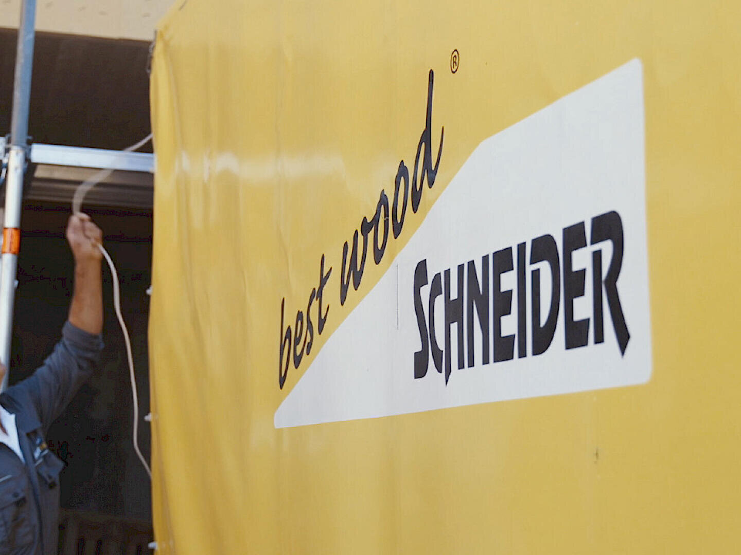 Eine Person zieht an einem Seil in der Nähe eines großen gelben Banners mit dem Text best wood SCHNEIDER in schwarzer und weißer Schrift. Nur der Arm der Person ist sichtbar.