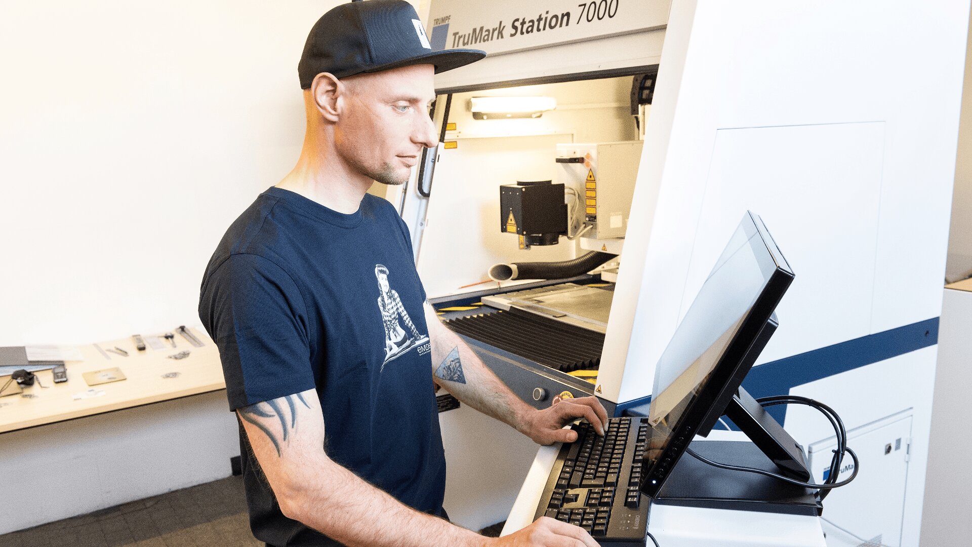 Ein Mann mit Mütze und marineblauem T-Shirt arbeitet an einem Computer vor einer großen Industriemaschine mit der Aufschrift TruMark Station 7000 in einer hellen, modernen Werkstatt.