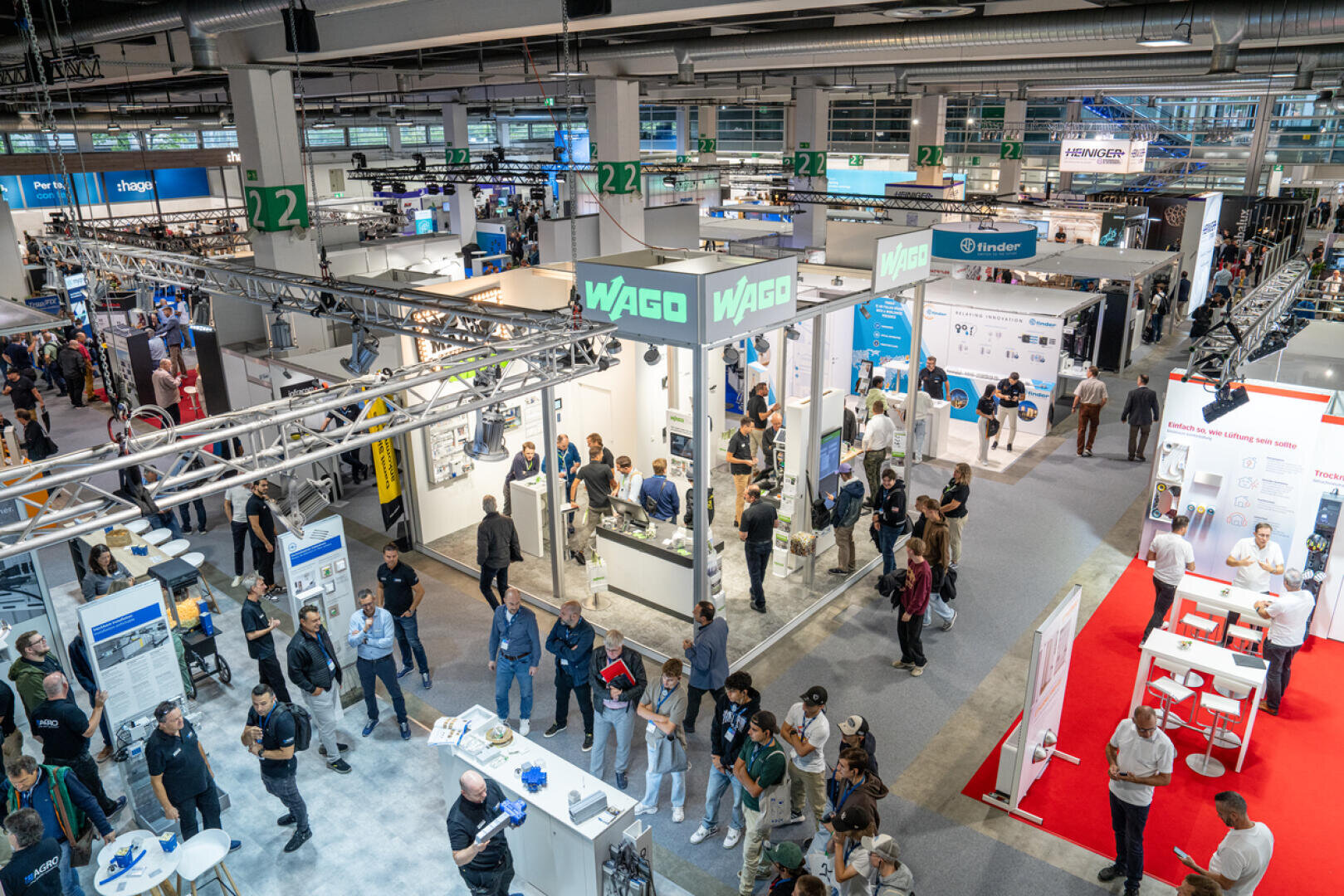 Eine belebte Messehalle mit zahlreichen Ausstellungsständen, darunter ein auffälliger WAGO-Stand, und vielen Besuchern, die unter heller Deckenbeleuchtung umhergehen, sich unterhalten und die Ausstellungsstücke erkunden.