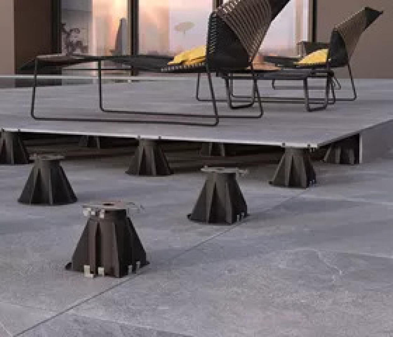 Außenterrasse mit erhöhten Fliesen, die von verstellbaren schwarzen Sockeln getragen werden; moderne Stühle und Kissen stehen auf der erhöhten Plattform, mit einem Gebäude und einem Sonnenuntergang im Hintergrund.