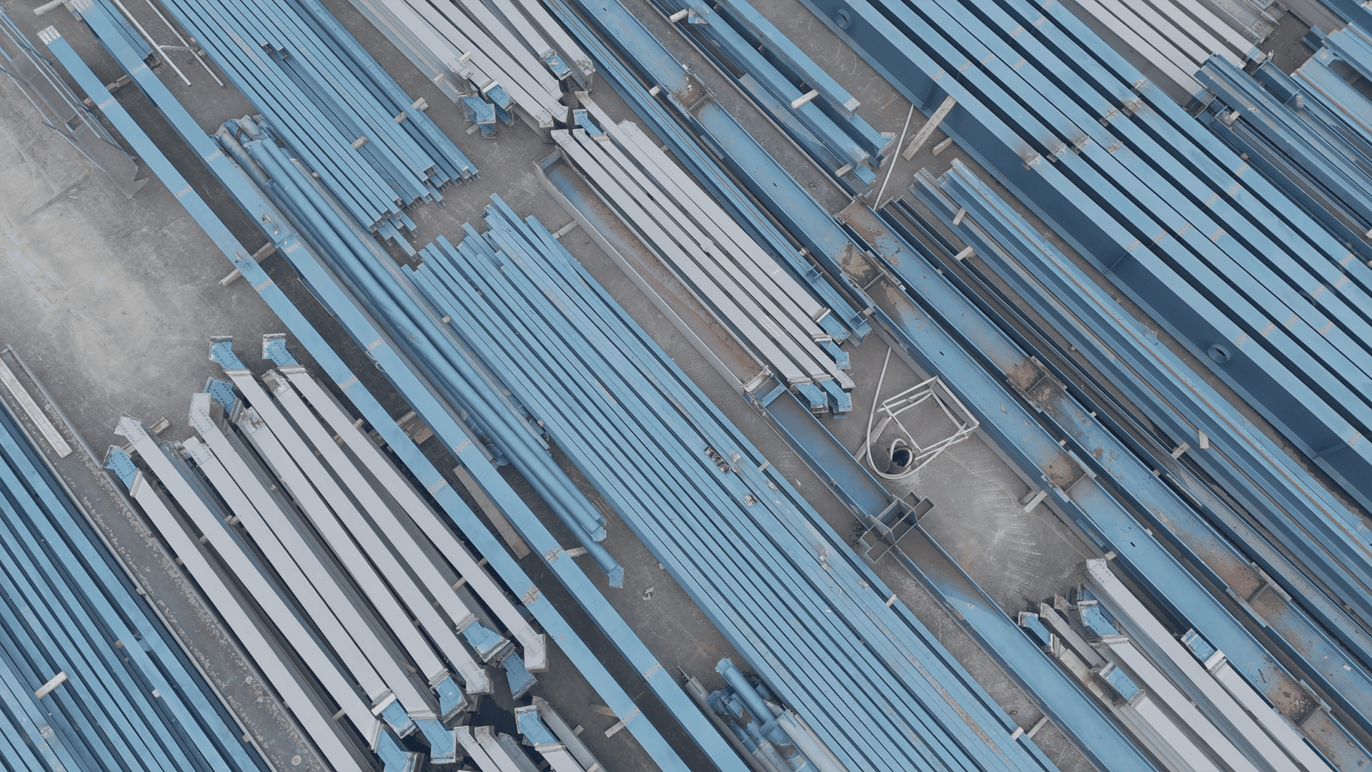 Vue aérienne de poutres d'acier bleues et blanches finement empilées, disposées parallèlement sur une surface en béton, peut-être sur un chantier de construction ou une aire de stockage.