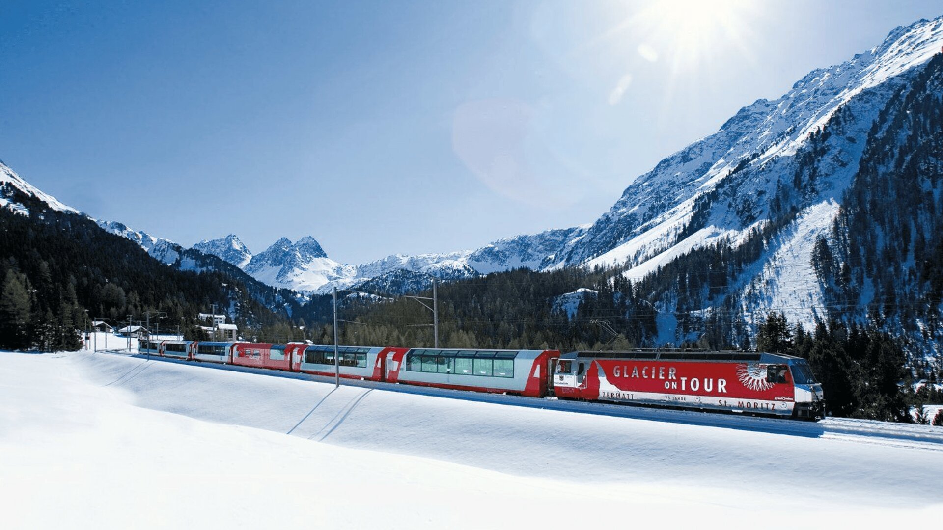 Ein roter Glacier Express-Zug fährt durch eine verschneite Alpenlandschaft, umgeben von hohen Tannen und schneebedeckten Bergen unter einem klaren blauen Himmel.