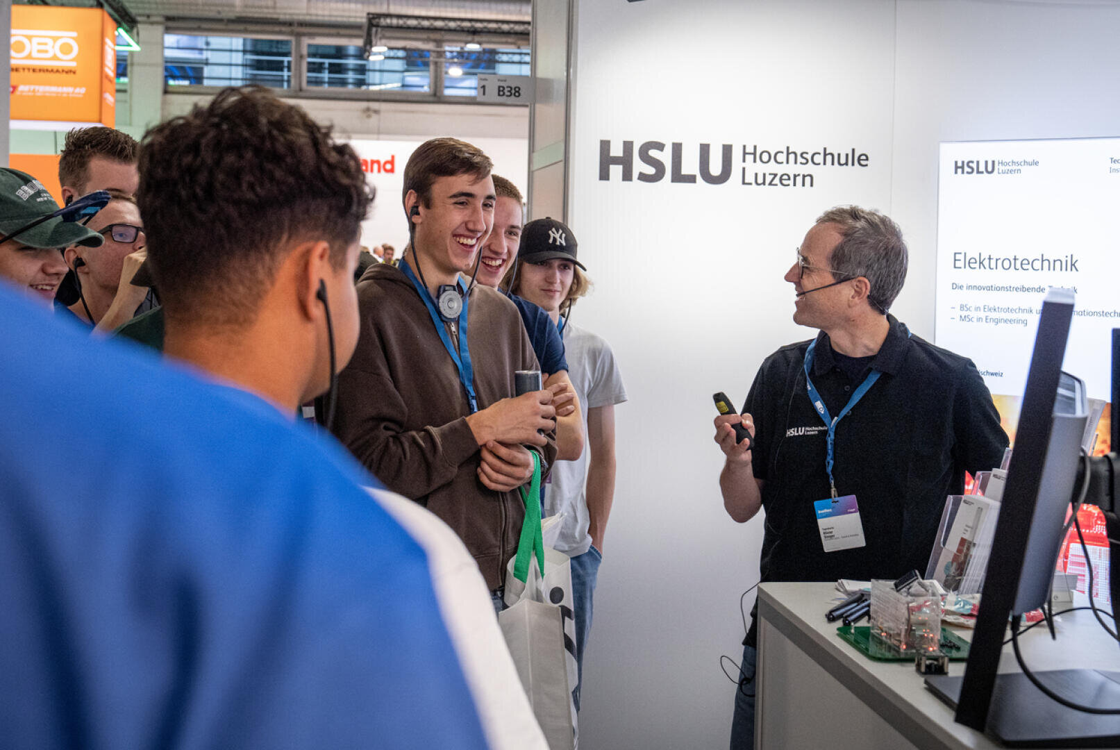 Eine Gruppe junger Menschen lächelt und unterhält sich mit einem Mann am Stand der HSLU (Hochschule Luzern) mit der Aufschrift „Elektrotechnik“ bei einer Indoor-Veranstaltung oder Messe. Der Mann scheint gerade etwas zu erklären.