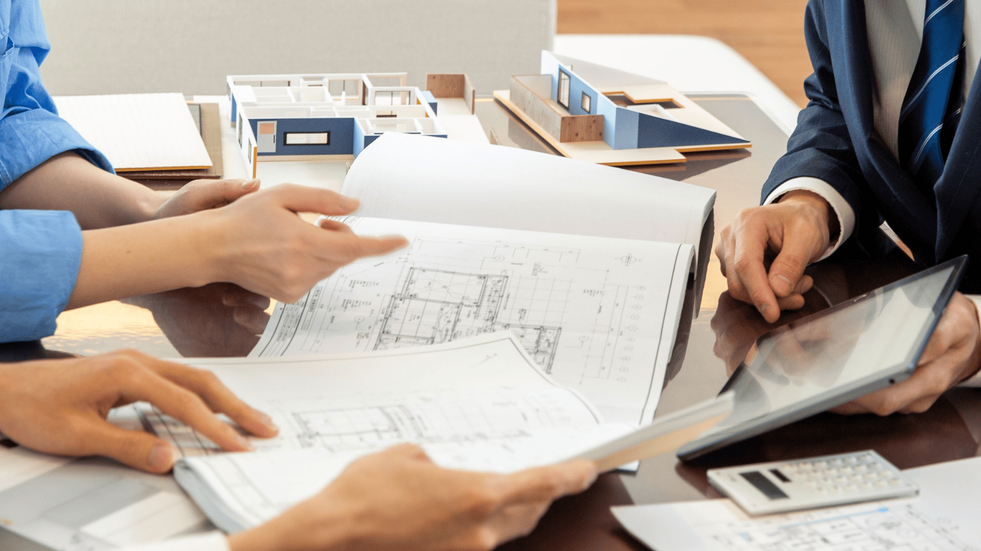 Trois personnes qui examinent des projets et des documents architecturaux à une table équipée de maquettes architecturales, d'une calculatrice et d'une tablette, ce qui indique une réunion ou une discussion sur la conception ou la planification de bâtiments.