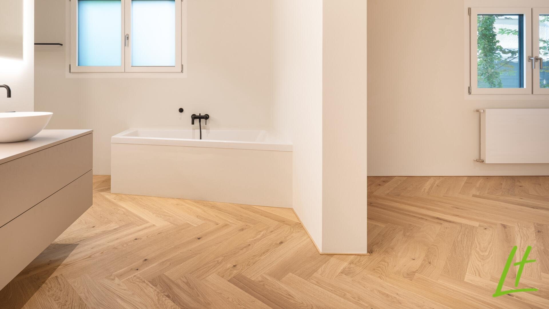 Minimalistisches Badezimmer mit hellem Fischgrätenholzboden, einer weißen Badewanne mit schwarzen Armaturen, einem modernen Waschbecken auf einem schwebenden Waschtisch und zwei Milchglasfenstern, die natürliches Licht hereinlassen.