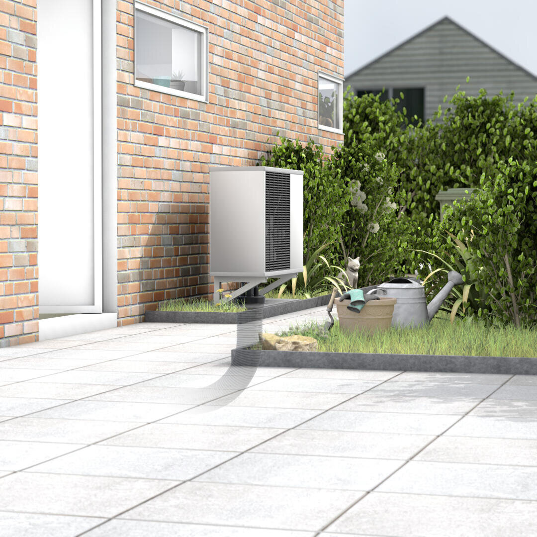 Une unité de pompe à chaleur montée à l'extérieur d'une maison en briques sur une terrasse, avec un arrosoir, des outils de jardinage et des plantes à proximité. En arrière-plan, on peut voir un bâtiment d'habitation et des buissons.