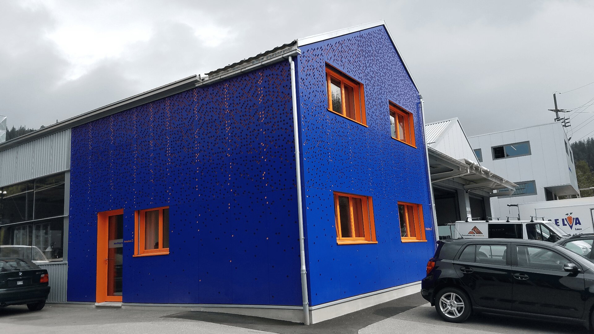 Ein modernes zweistöckiges Gebäude mit leuchtend blauem Äußeren und orange gerahmten Fenstern steht neben geparkten Autos. Der Himmel ist bedeckt, und in der Nähe befinden sich weitere Gebäude im Industriestil.