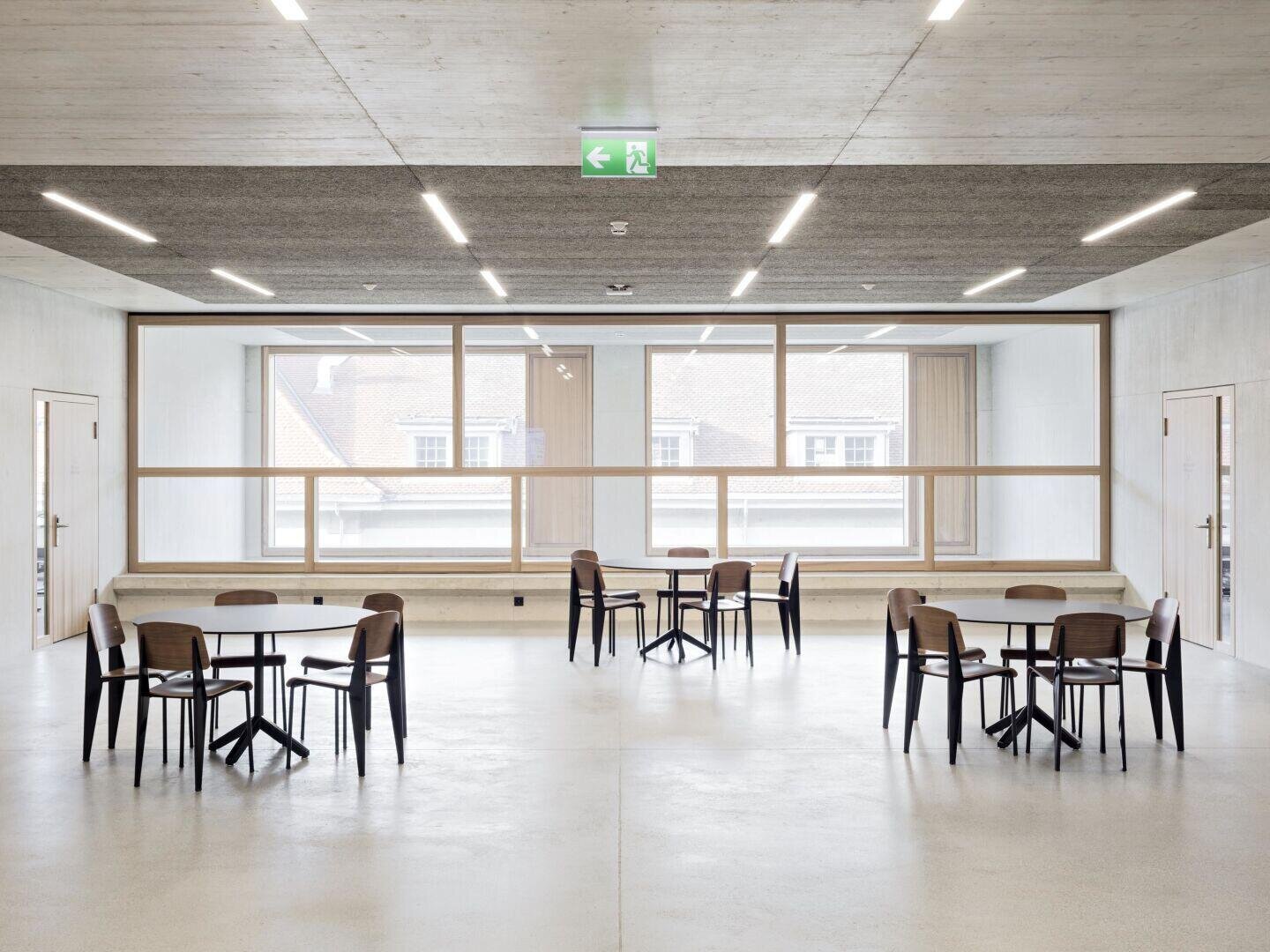 Une pièce spacieuse et moderne avec quatre tables et plusieurs chaises, de grandes fenêtres qui laissent entrer la lumière naturelle, des murs et des sols clairs et un panneau de sortie de secours au plafond.