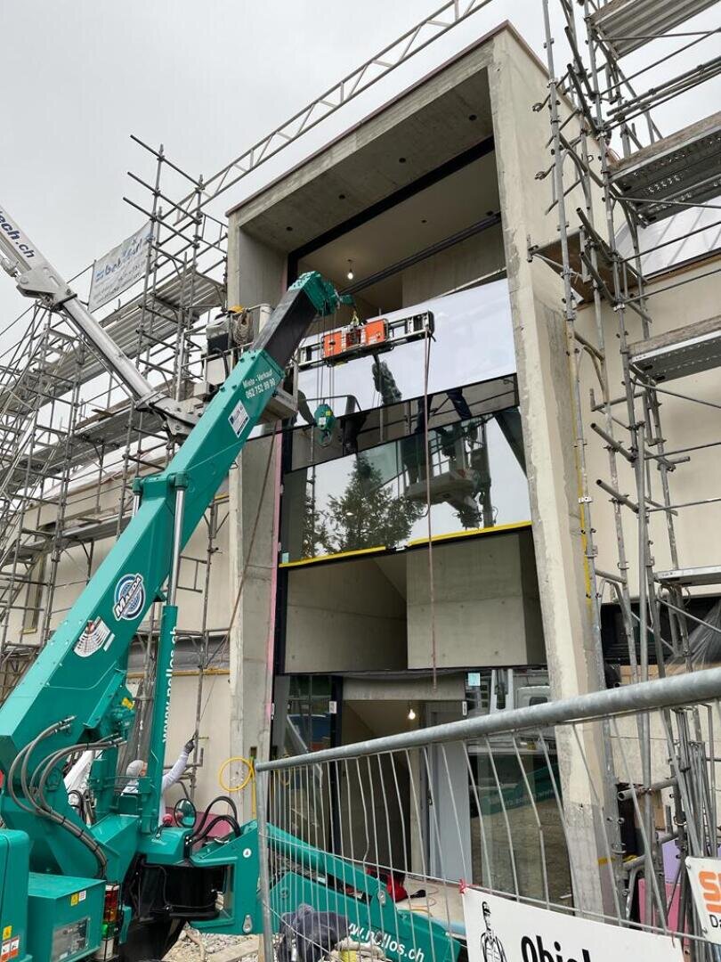 Eine Baustelle mit Gerüsten umgibt ein modernes Gebäude. Arbeiter an einem grünen Kranaufzug installieren ein großes Glasfenster im zweiten Stock. Im Vordergrund sind Sicherheitsbarrieren und Geräte zu sehen.