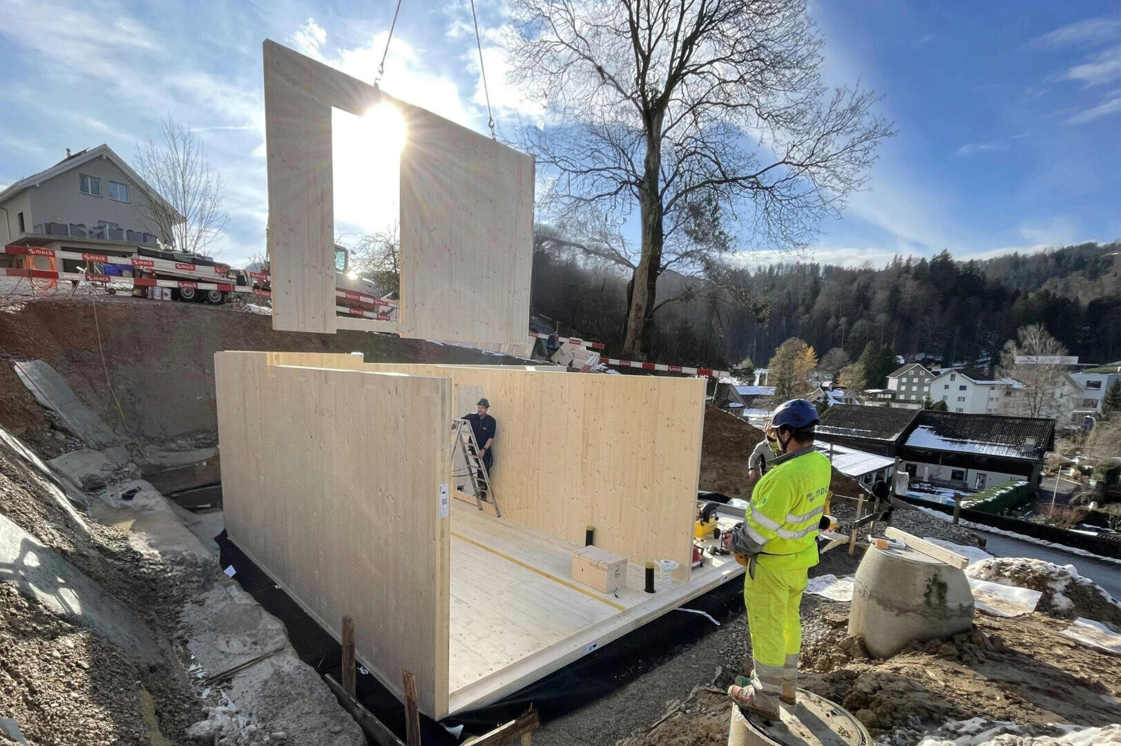 Eine Baustelle, auf der Arbeiter ein hölzernes Gebäude zusammensetzen; ein Kran hebt eine große hölzerne Wandplatte über den Kopf, während ein Arbeiter in leuchtend gelber Kleidung zusieht. Im Hintergrund sind Häuser, Bäume und ein blauer Himmel zu sehen.