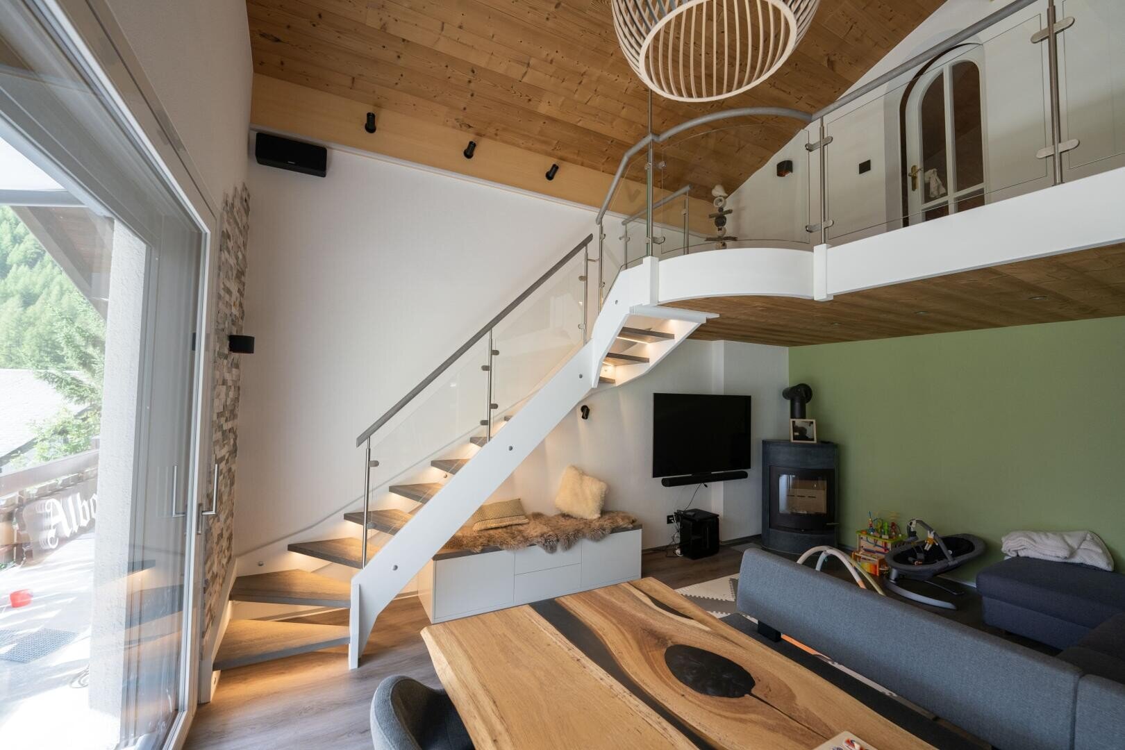 Modernes Wohnzimmer mit Holzdecke, Glastreppe, Esstisch aus hellem Holz, TV, Kamin, Kinderspielzeug und Blick auf einen Balkon. Natürliches Licht füllt den Raum und unterstreicht die moderne Einrichtung.