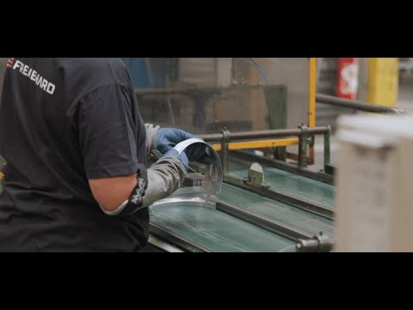 Ein Arbeiter mit Handschuhen hantiert mit einem durchsichtigen Kunststoffgesichtsschutz an einem Förderband in einer Industrieumgebung.