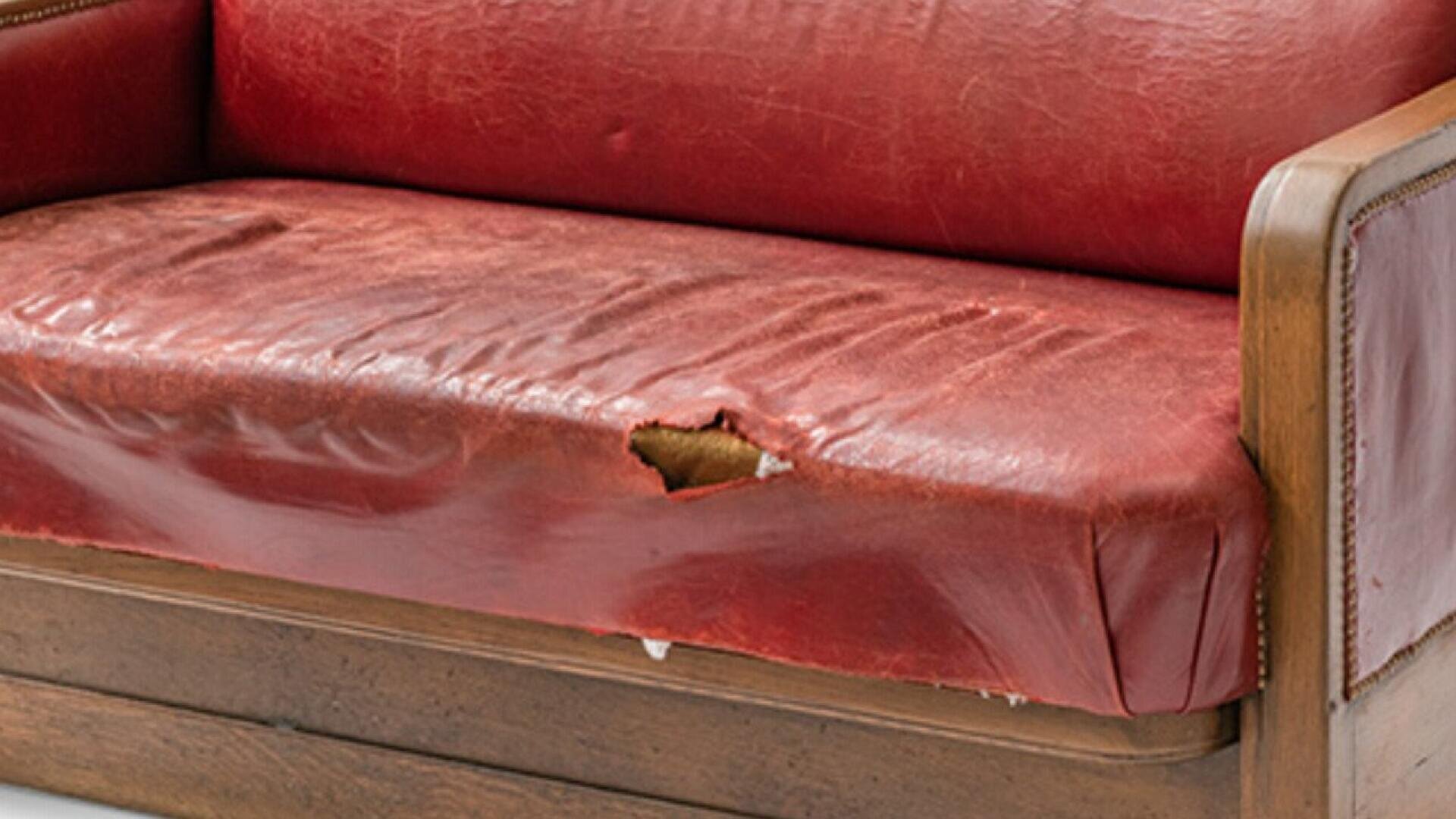Gros plan sur un canapé en cuir rouge usé, dont le coussin d'assise est largement déchiré, laissant apparaître le tissu sous-jacent. Les accoudoirs et le piètement sont en bois.
