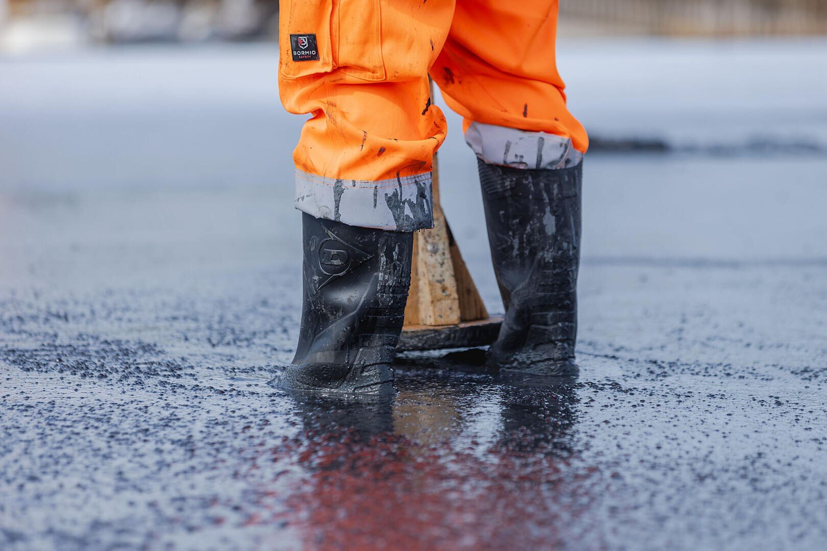 Une personne portant un pantalon orange imperméable et des bottes en caoutchouc noires se tient dans une eau peu profonde, le bas des jambes et les pieds étant visibles. La surface de l'eau se reflète et semble froide, peut-être glacée.