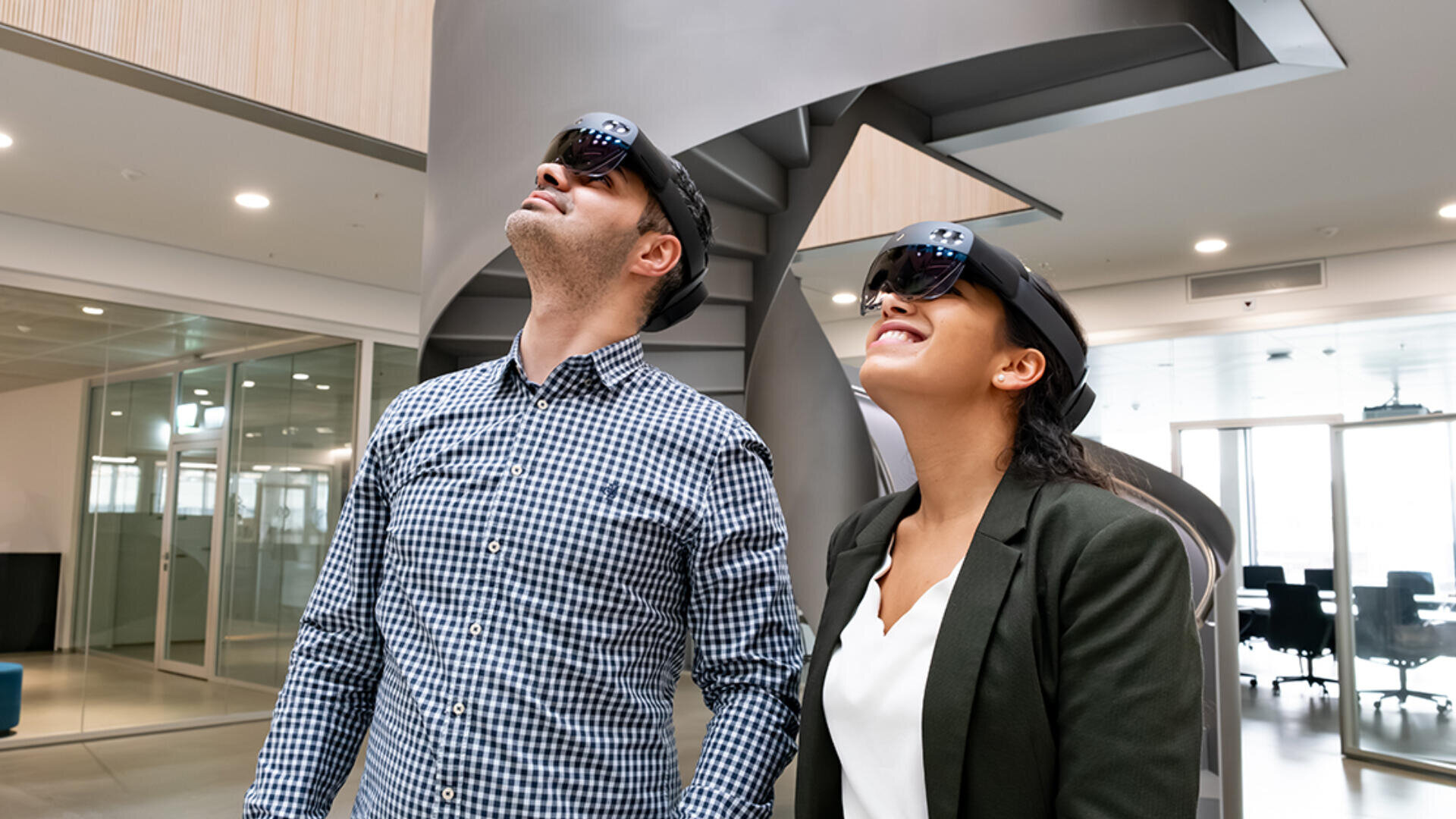 Zwei Personen, die Augmented-Reality-Headsets tragen, stehen in einer modernen Büroumgebung mit Glaswänden und einer Wendeltreppe hinter ihnen, schauen nach oben und lächeln.