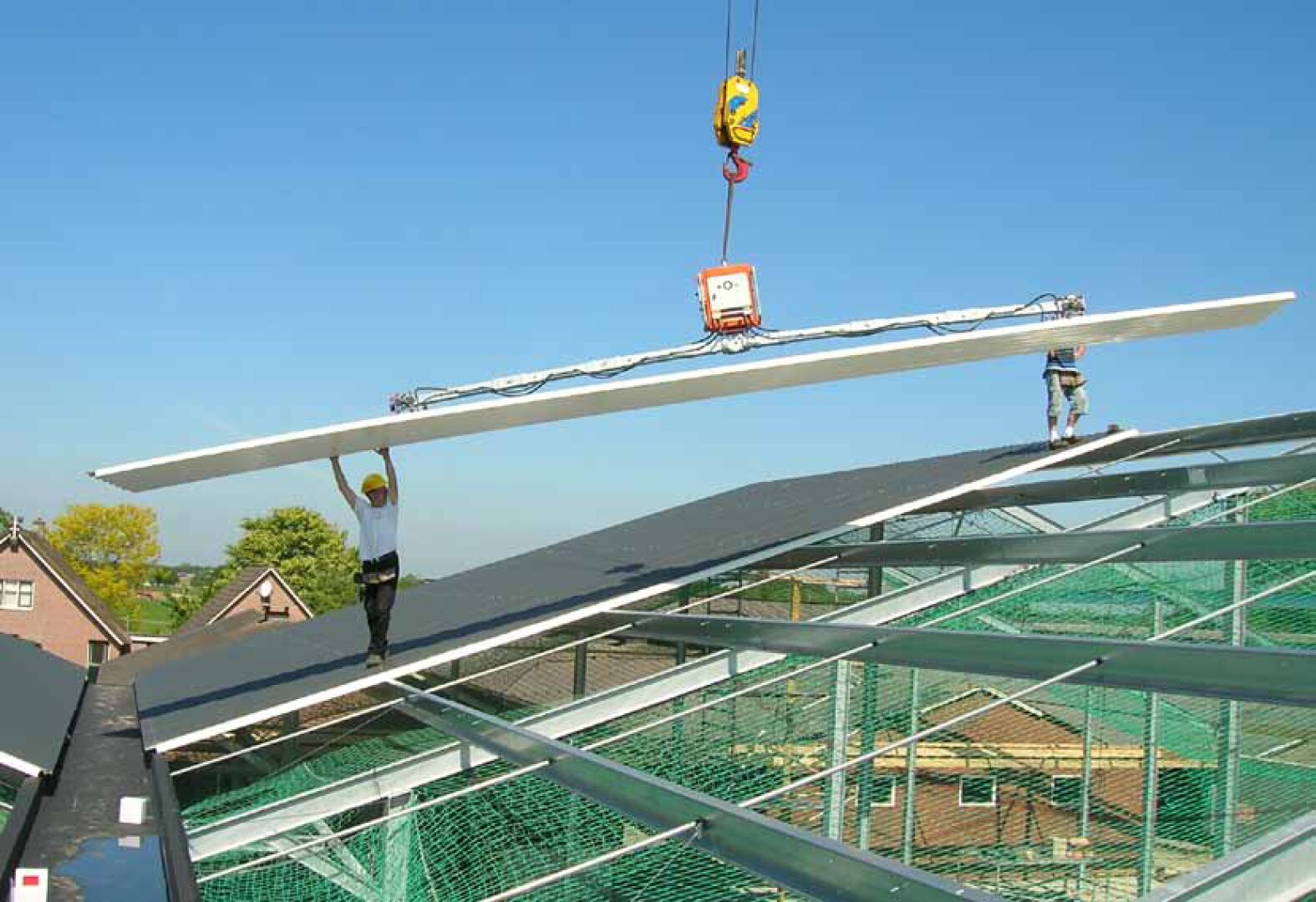 Zwei Bauarbeiter stehen auf einem schrägen Metalldachstuhl, während ein Kran unter blauem Himmel eine große, flache Bauplatte an ihren Platz hebt. Unter ihnen sind Sicherheitsnetze zu sehen.