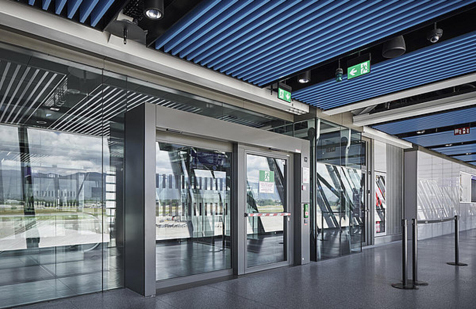 Automatische Glastüren in einem Flughafenterminal, mit blauer Lamellendecke, Notausgangsschildern darüber, Spiegelungen in den Glaswänden und einer Seilabsperrung in der Nähe. Natürliches Licht scheint durch große Fenster.