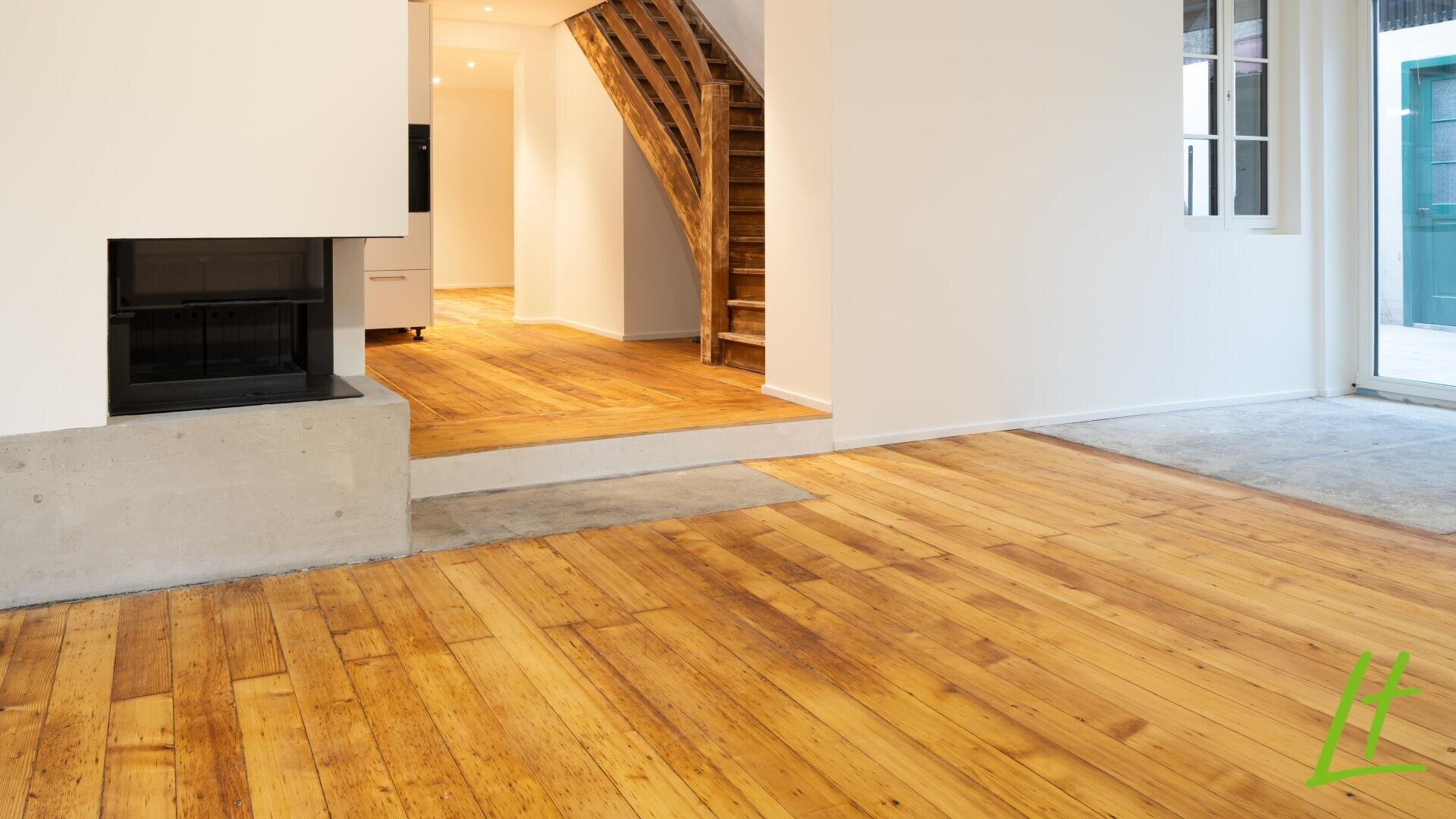 Un intérieur lumineux avec des sols en bois, une cheminée sur la gauche, un escalier en bois au milieu, des murs blancs et de grandes fenêtres qui laissent entrer la lumière naturelle. Un escalier en béton sépare deux parties de l'espace de vie.