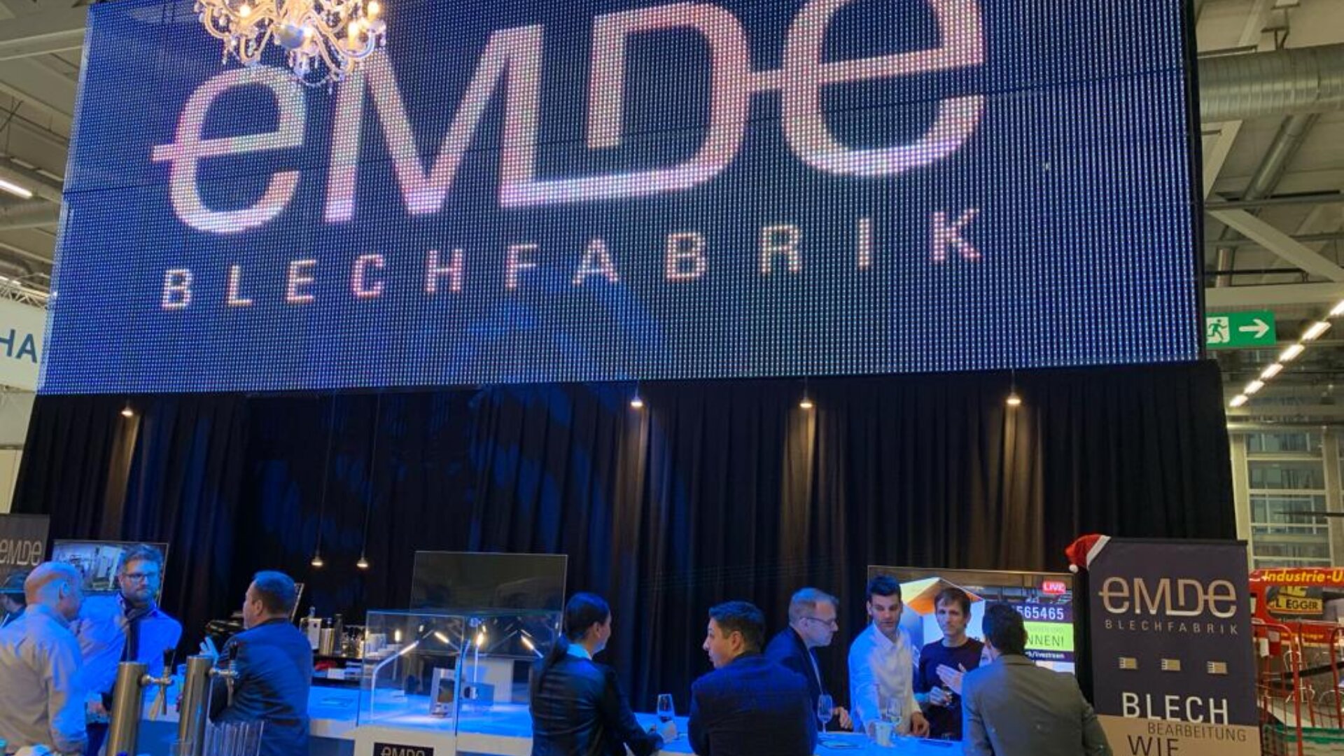 Eine Gruppe von Menschen steht und unterhält sich an einem Ausstellungsstand unter einem großen digitalen Schild mit der Aufschrift eMDE BLECHFABRIK in einem hell erleuchteten Veranstaltungsraum.