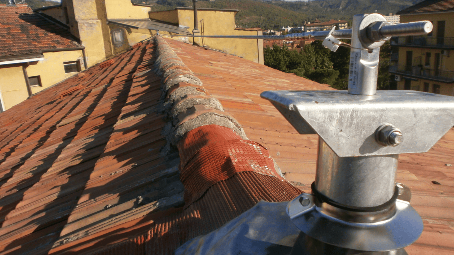 Eine Metallantenne ist auf einem roten Ziegeldach angebracht, dessen First mit einem Schutznetz bedeckt ist. Im Hintergrund sind bei klarem Himmel gelbe Gebäude und grüne Hügel zu sehen.