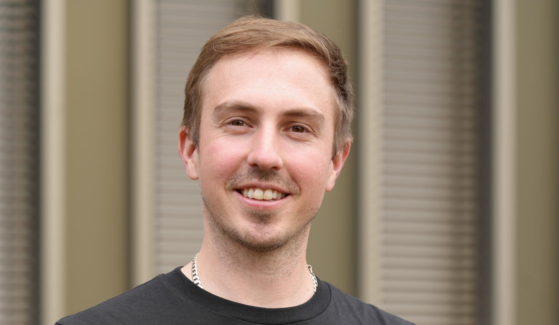 Ein junger Mann mit kurzen hellbraunen Haaren und einem gestutzten Bart lächelt in die Kamera. Er trägt ein schwarzes T-Shirt und eine Kettenhalskette und steht vor einem Hintergrund mit unscharfen vertikalen Flächen.