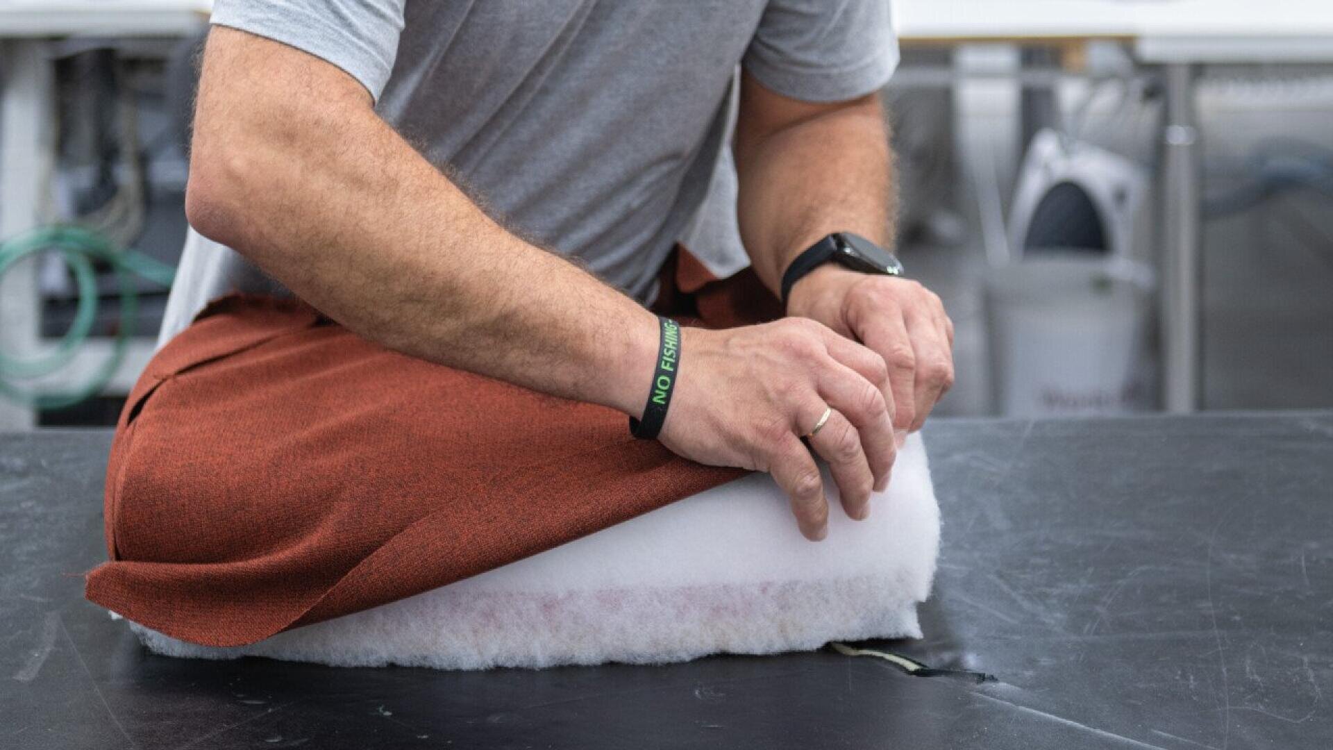 Une personne portant une chemise grise et des bracelets enroule un tissu orange autour d'un coussin blanc, travaillant sur le rembourrage d'une table noire.