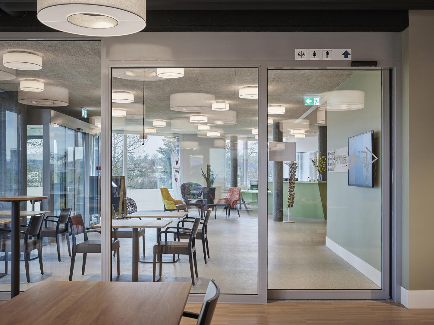 Coin salon moderne à l'intérieur avec vue à travers des portes vitrées, chaises colorées, tables et grandes fenêtres qui laissent entrer la lumière naturelle ; des plafonniers et des panneaux muraux sont également visibles.