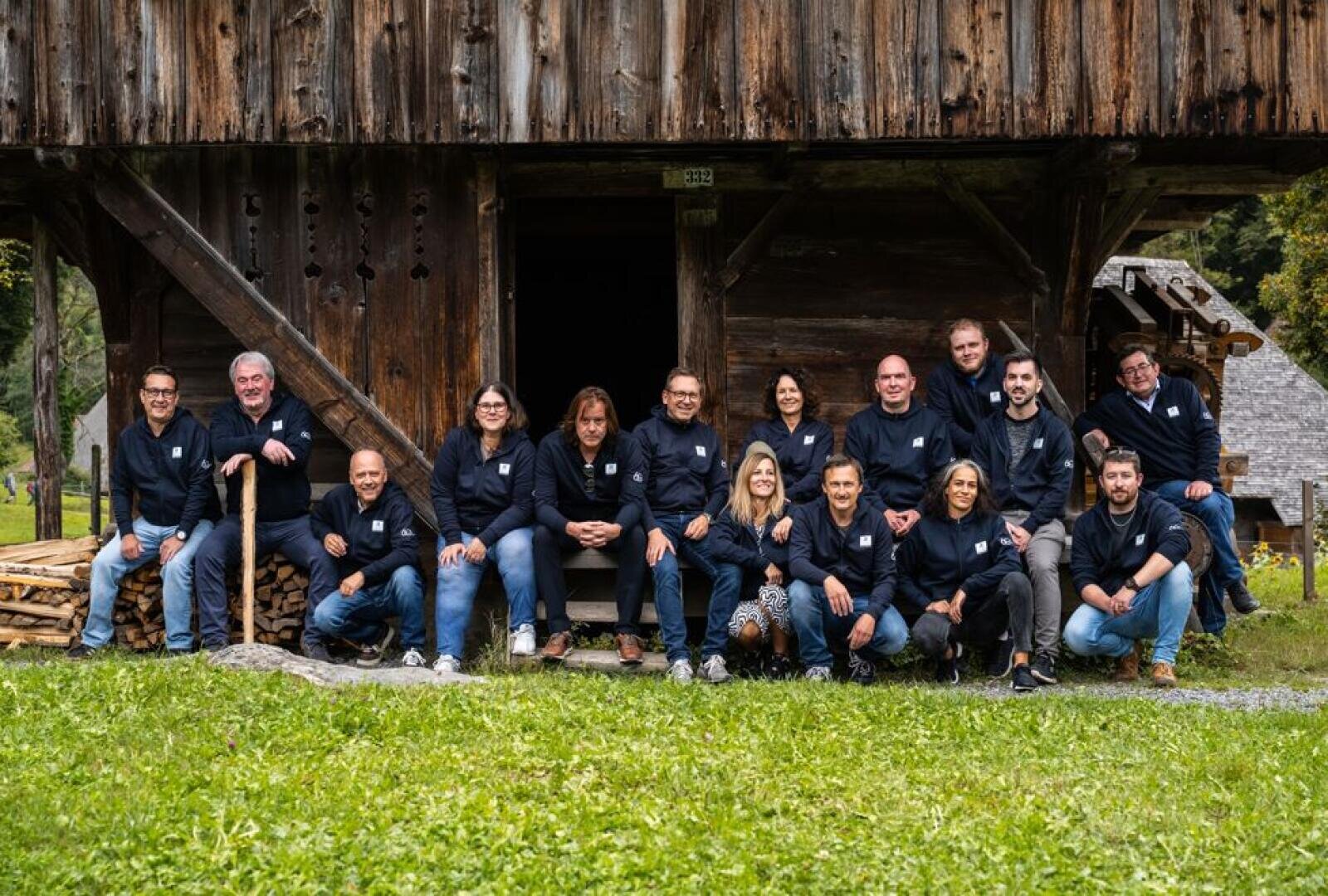 Un groupe de seize personnes portant des vestes sombres assorties posent ensemble devant un bâtiment rustique en bois, assises et debout sur des marches et de l'herbe, avec du bois de chauffage empilé à proximité.