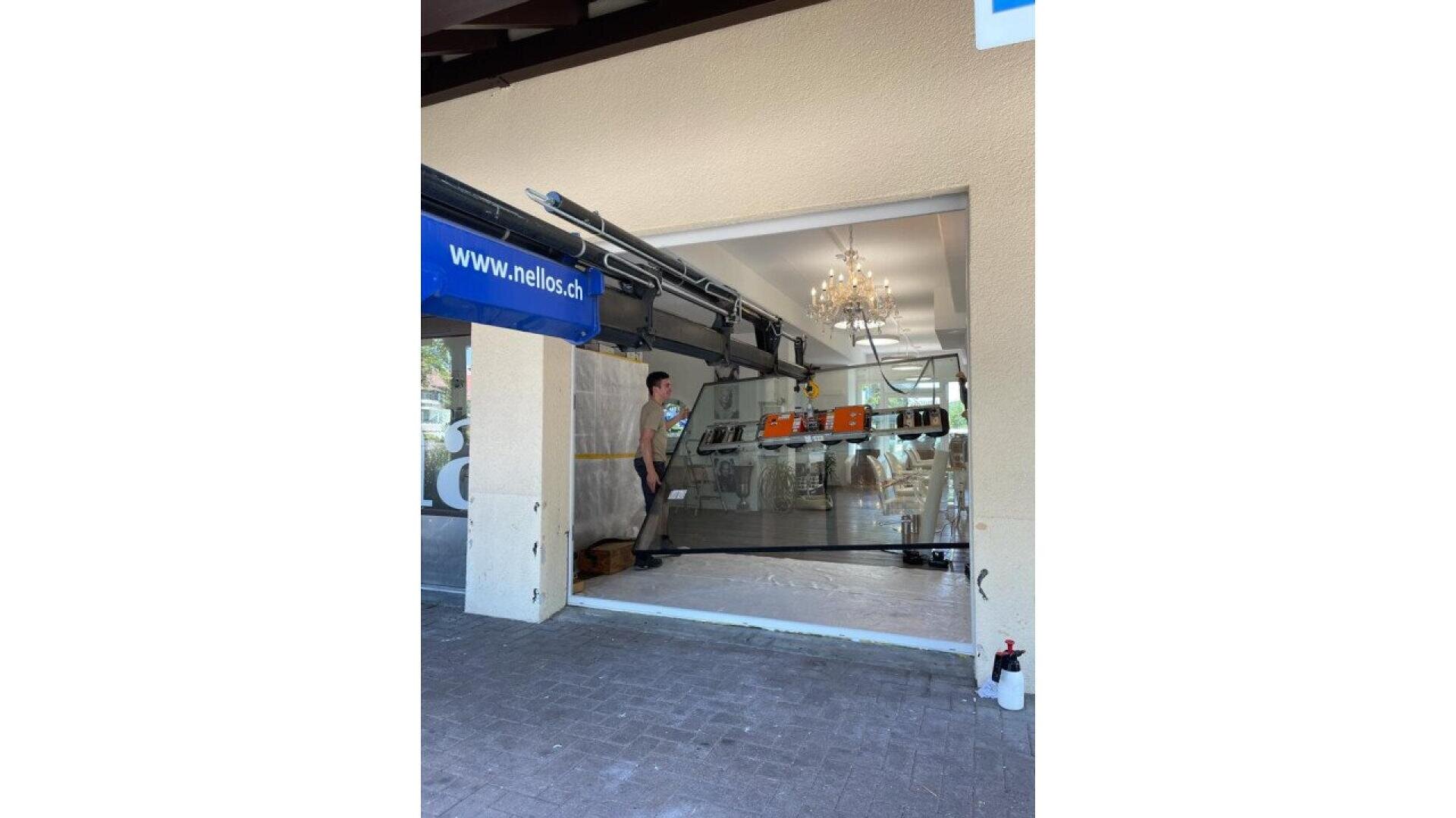 Une personne se tient près de la porte vitrée automatique partiellement ouverte d'un restaurant ou d'un café. À l'intérieur, des tables et des chaises sont disposées et un lustre est suspendu au plafond. La porte est munie d'un panneau bleu sur lequel on peut lire "nello.ch".
