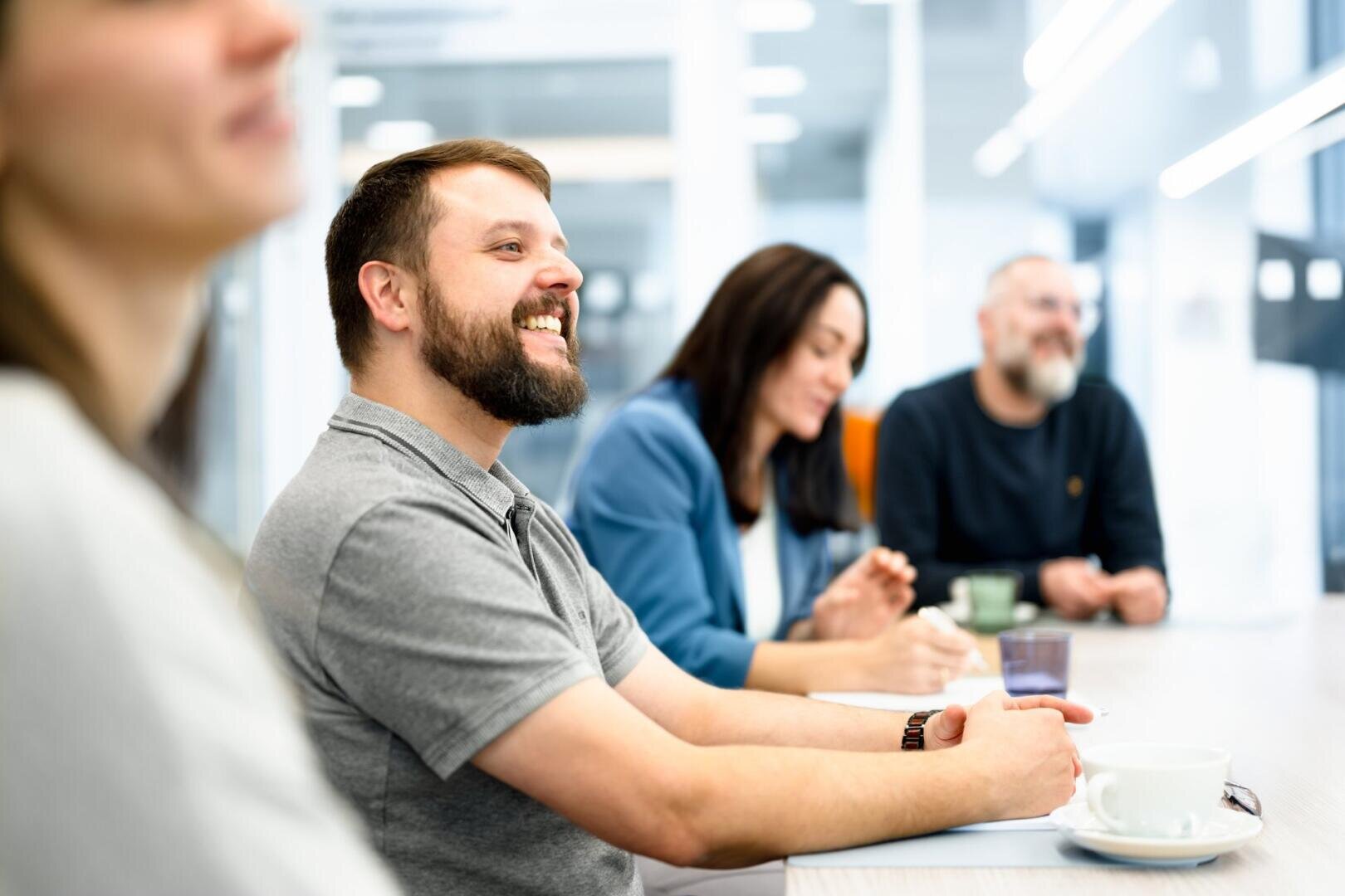 Un groupe de quatre adultes est assis à une table dans un bureau moderne et lumineux, souriant et discutant pendant une réunion. Un homme au premier plan rit, tandis que les autres semblent attentifs et détendus.