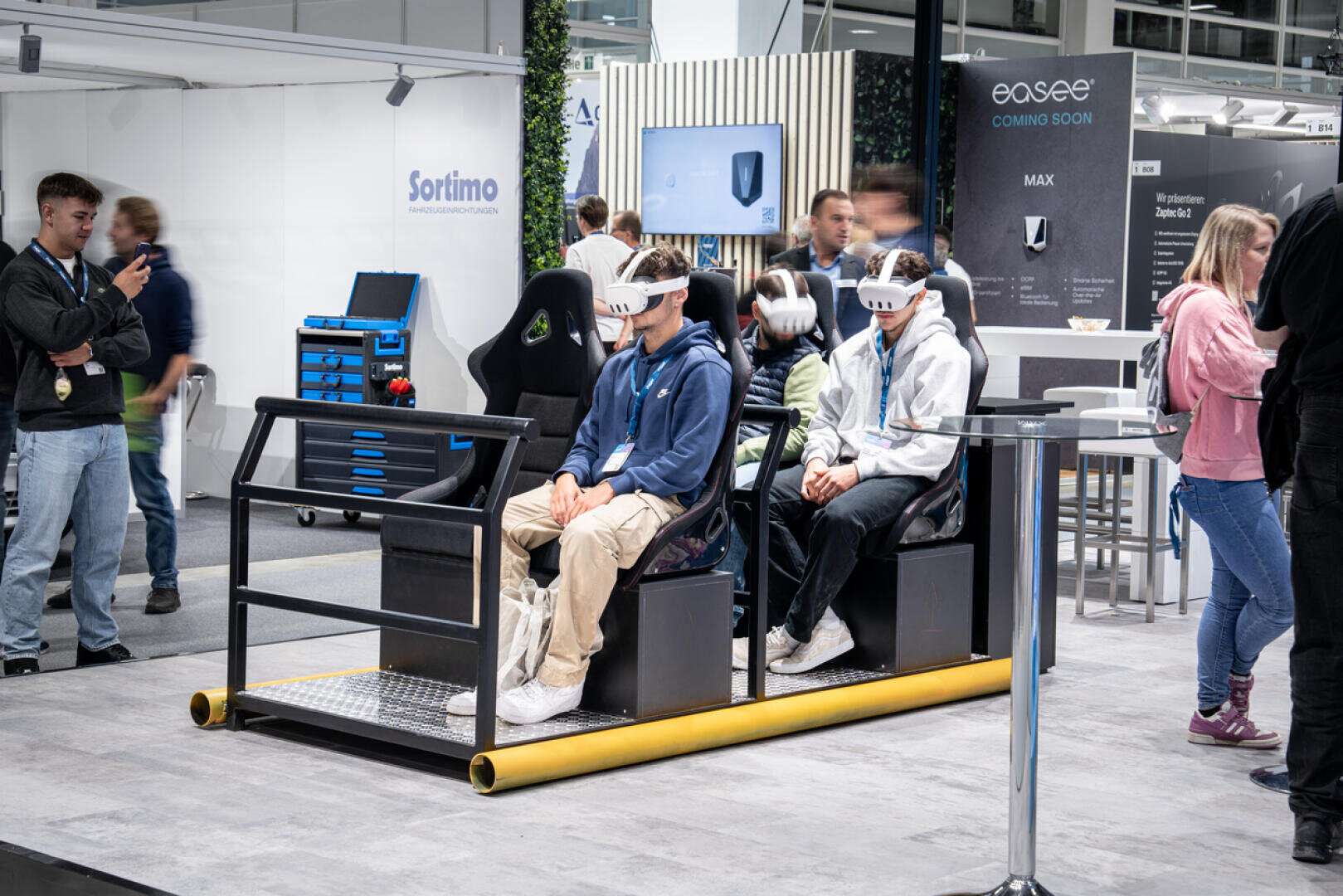 Vier Personen sitzen mit VR-Headsets in einem Bewegungssimulator auf einer Technologiemesse, während andere herumstehen und zusehen. Im Hintergrund sind Stände und Technologieausstellungen zu sehen.
