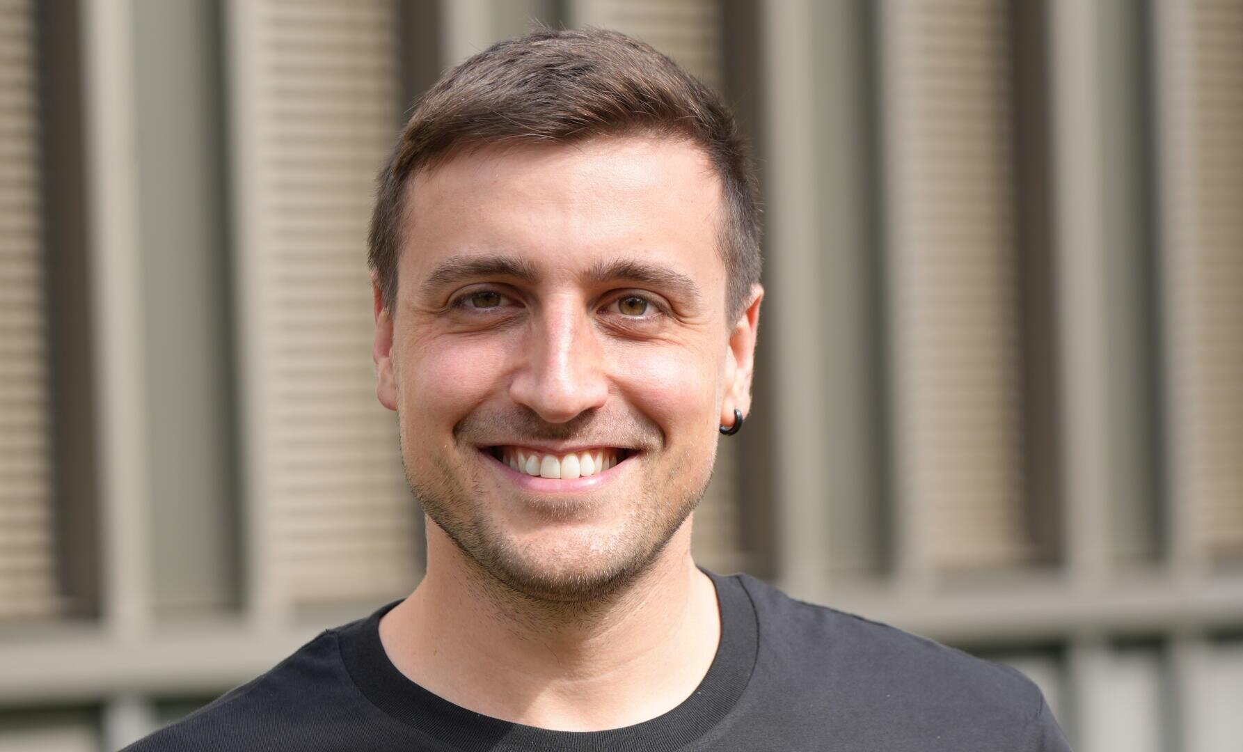 Ein Mann mit kurzen braunen Haaren und einem Ohrring lächelt in die Kamera, er trägt ein schwarzes Hemd. Der Hintergrund ist mit vertikalen Linien und Fensterläden verschwommen.