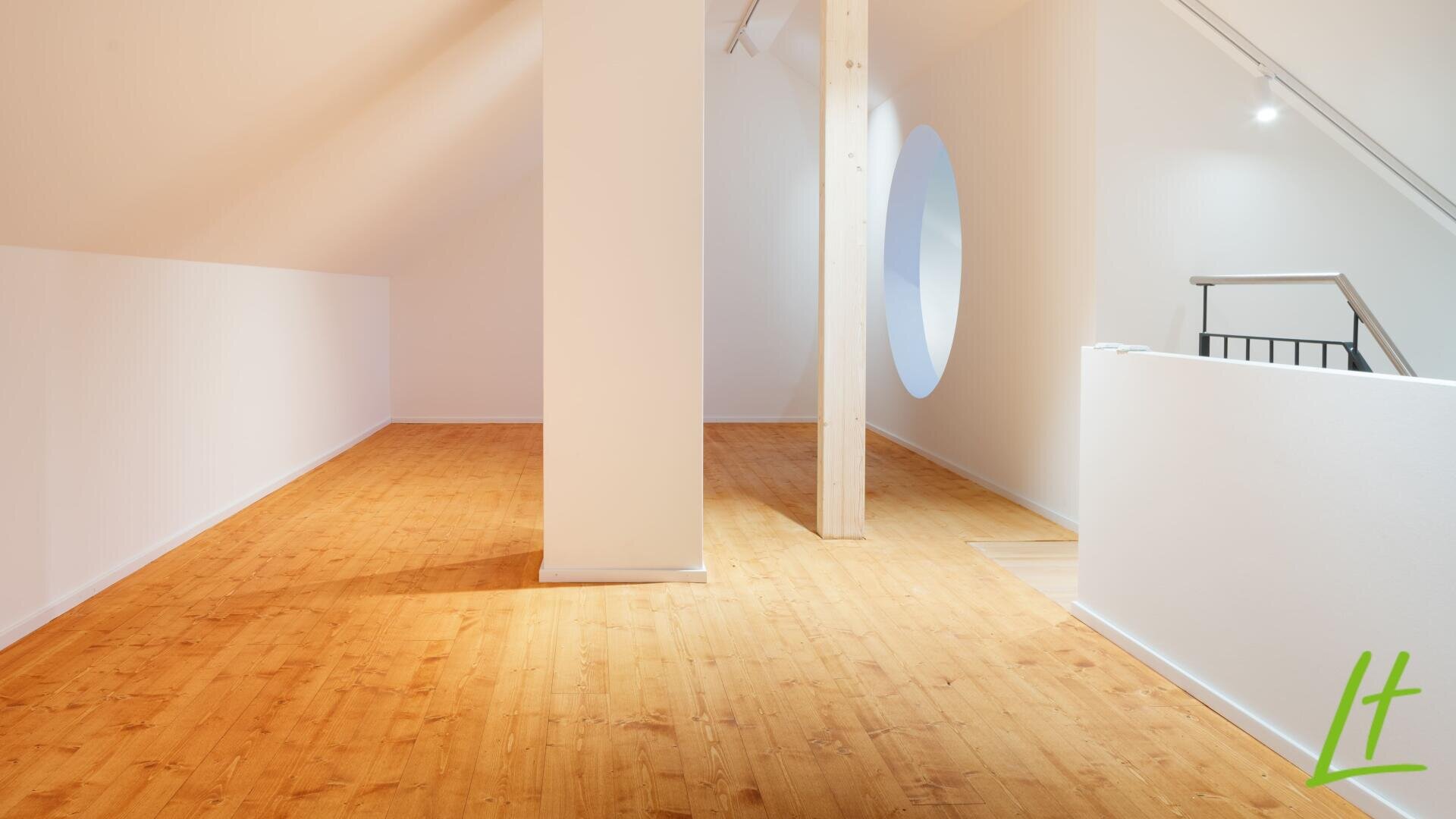 Une pièce loft lumineuse et moderne avec un sol en bois clair, des murs blancs, une colonne de soutien centrale, une haute poutre en bois, une fenêtre ronde et une balustrade minimaliste sur le côté droit. Le logo vert "Lt" apparaît dans le coin inférieur droit.