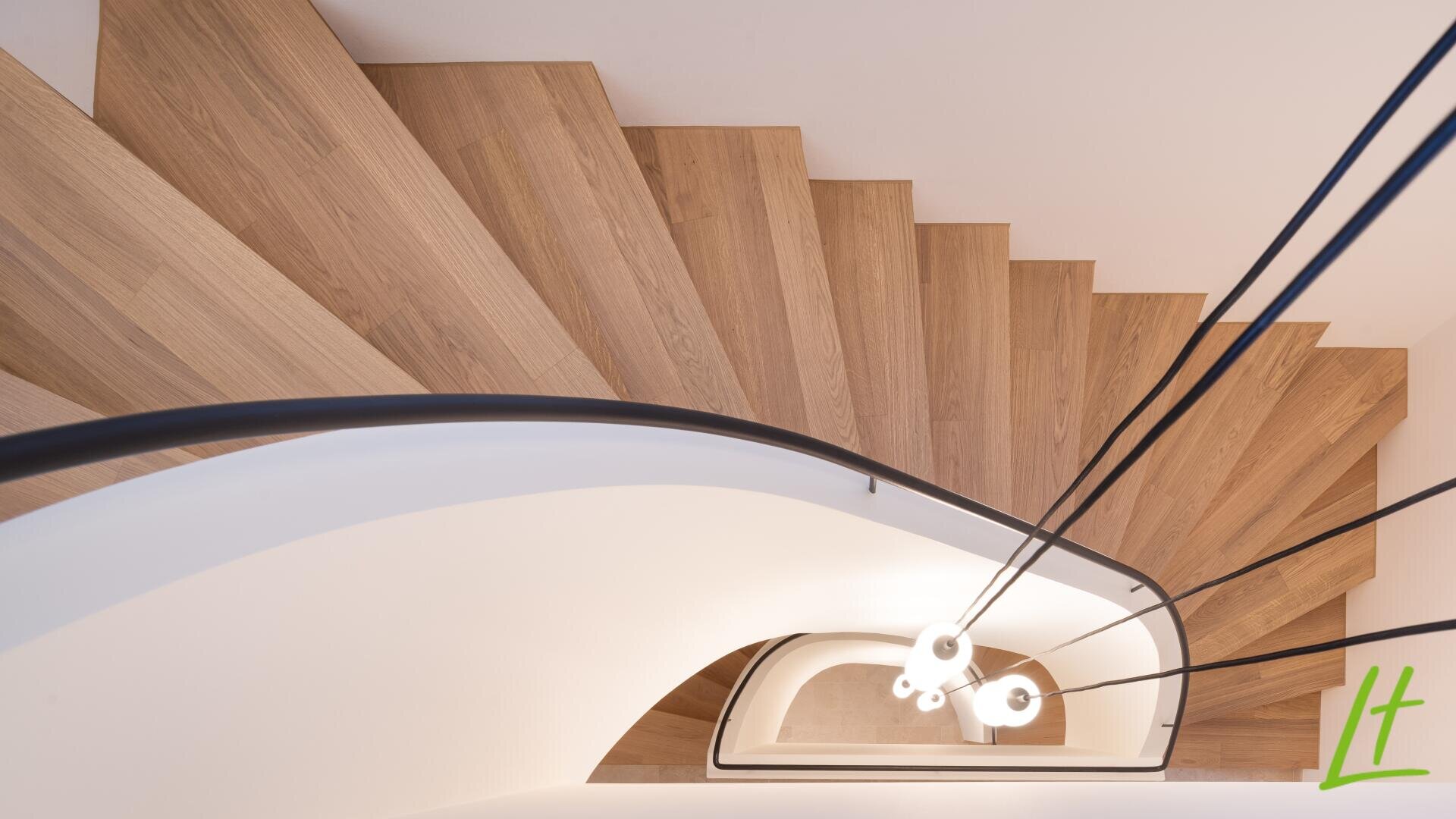 Blick von oben auf eine moderne Holzspindeltreppe mit schwarzem Geländer, weißen Wänden und runden Deckenleuchten in der Mitte.