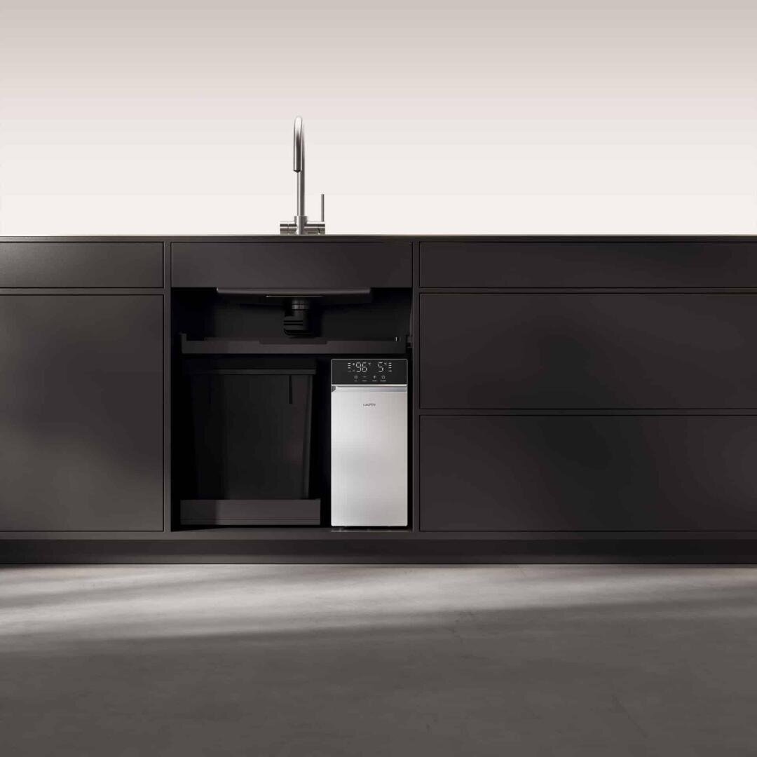 Des armoires de cuisine noires minimalistes surmontées d'un évier moderne. Un appareil compact argenté à affichage numérique est installé sous l'évier, entouré de placards vides. Le sol est sobre et gris.