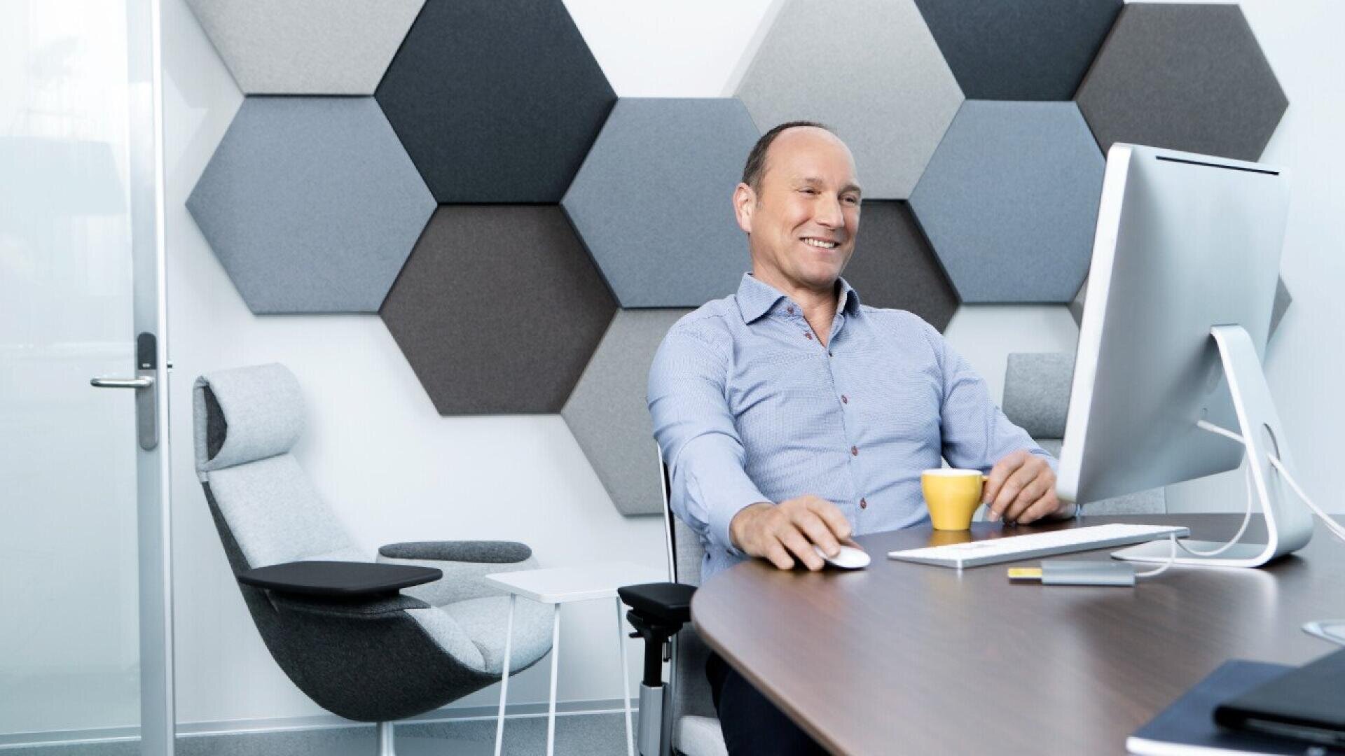 Un homme en chemise bleue est assis à un bureau, souriant, utilisant un ordinateur, avec une tasse de café jaune à proximité. Derrière lui se trouvent des chaises modernes grises et un mur décoré de grands panneaux hexagonaux gris.