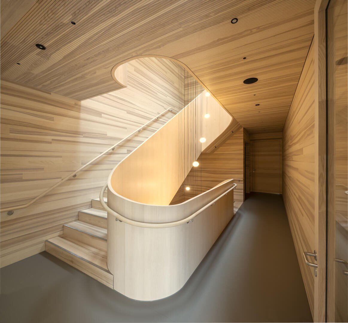 Eine moderne Treppe mit geschwungenen Holzwänden und einer geschwungenen Decke mit horizontalen Holzmaserungen, minimalistischer Beleuchtung und einem glatten Handlauf, die einen warmen und eleganten architektonischen Raum schaffen.