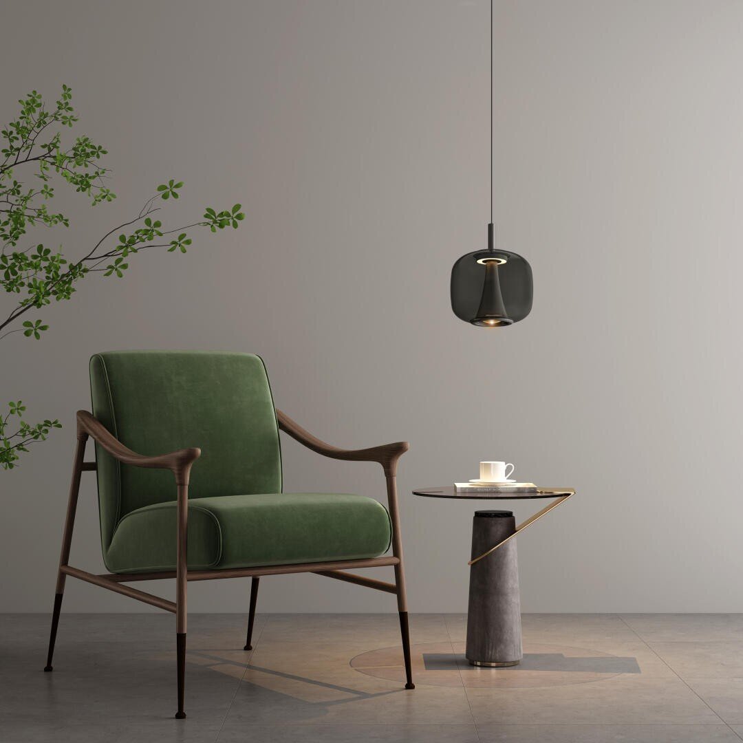 Un intérieur moderne avec un fauteuil vert, une petite table d'appoint ronde avec une tasse et une soucoupe, une suspension noire au-dessus et une plante à feuilles devant un mur gris sobre.