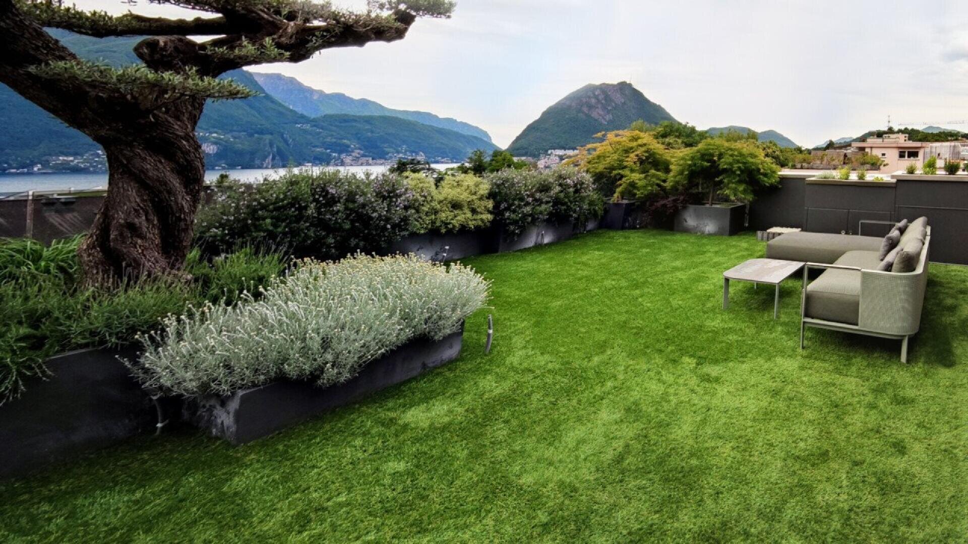 Un jardin sur le toit avec une pelouse verdoyante, des arbustes, un grand arbre, des sièges modernes en plein air et des vues panoramiques sur les montagnes et le lac en arrière-plan.