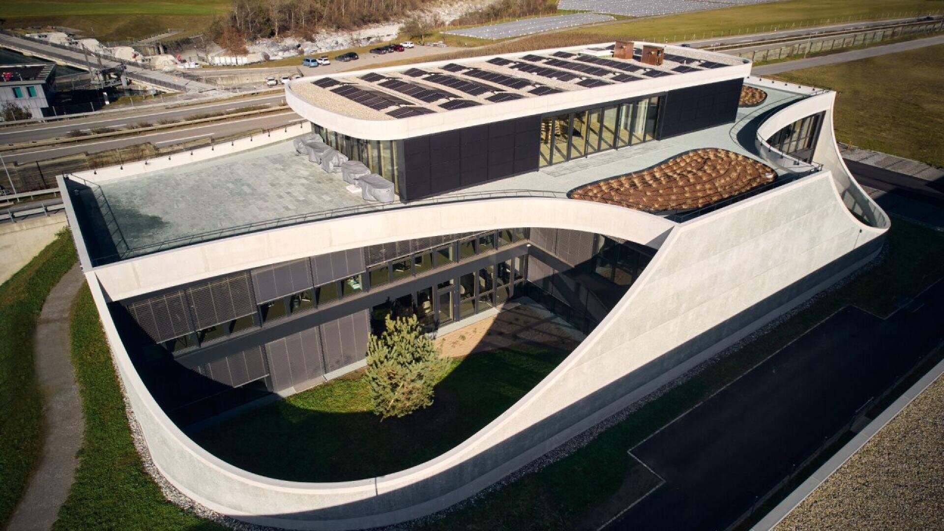 Vue aérienne d'un bâtiment moderne et incurvé, avec un extérieur blanc ondulé, des panneaux solaires sur le toit et des espaces verts. La structure est entourée d'herbe, de routes et de champs.