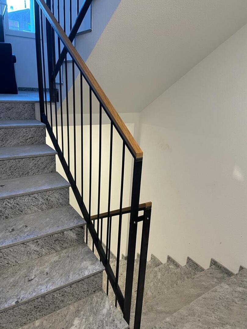 Ein Treppenhaus mit grauen Steinstufen, schwarzem Metallgeländer mit Holzhandlauf und weißen Wänden. Die Treppe biegt an einem Treppenabsatz ab und führt sowohl nach oben als auch nach unten. Durch ein nahe gelegenes Fenster scheint natürliches Licht herein.